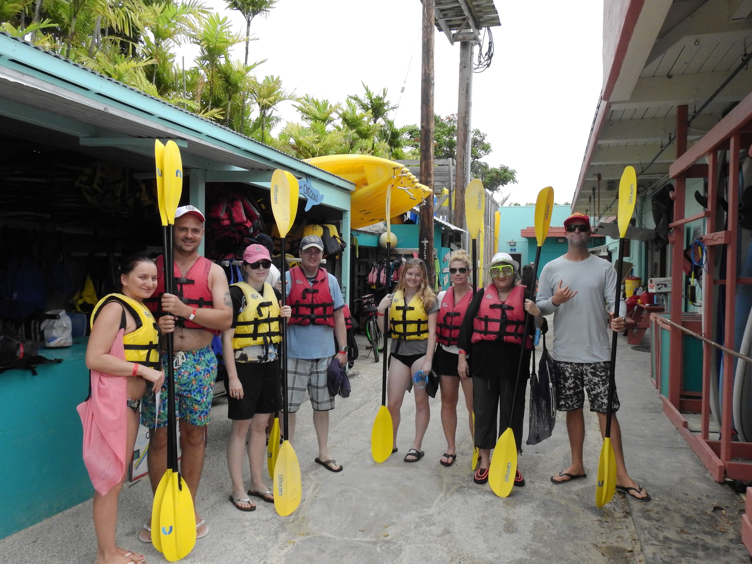 A group of nine people standing in a line, holding yellow paddles, ready for a kayaking activity, at Kailua Beach Adventures, wearing life jackets and casual beach attire.