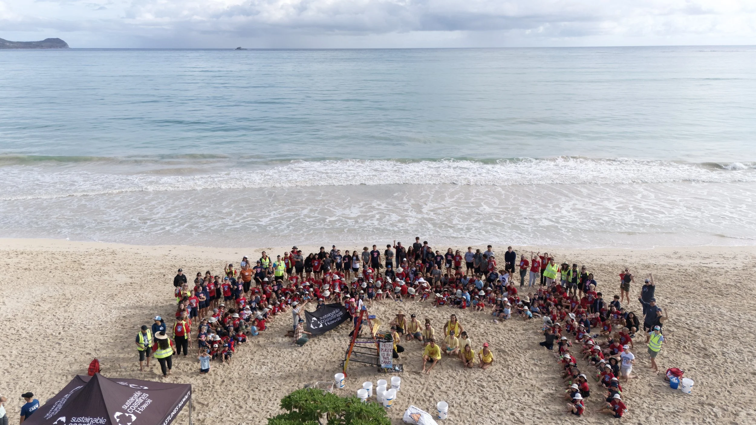 Large group of people, including children and adults, gathered on a sandy beach. They are arranged in the shape of a heart, with some holding signs and banners promoting sustainability and environmental awareness. The ocean with gentle waves and a cl