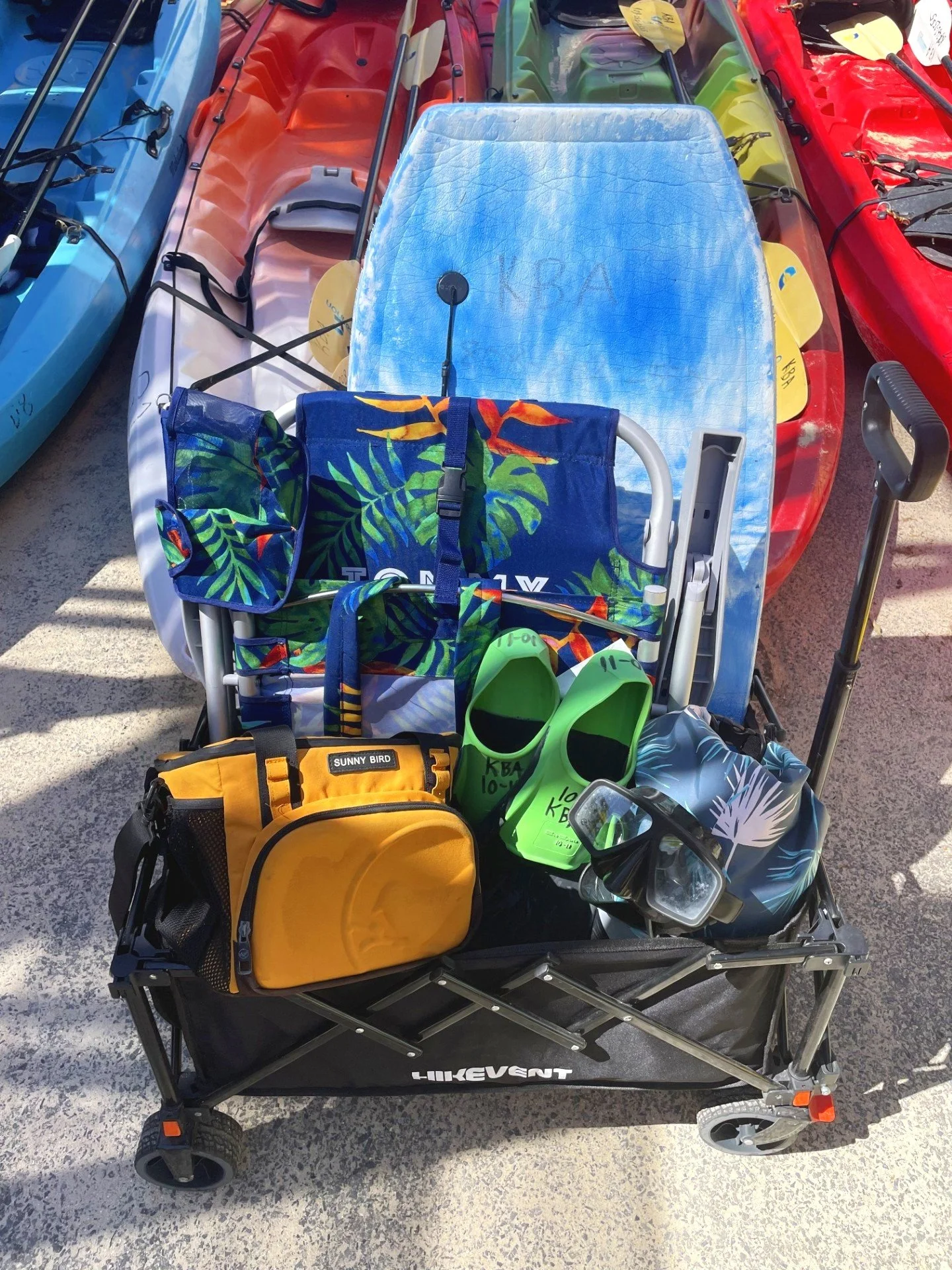 A cart filled with gear for kayaking, including a blue and green tropical-themed towel, a patterned dry bag, and a large blue towel. Kayaks are visible in the background. available for rental!