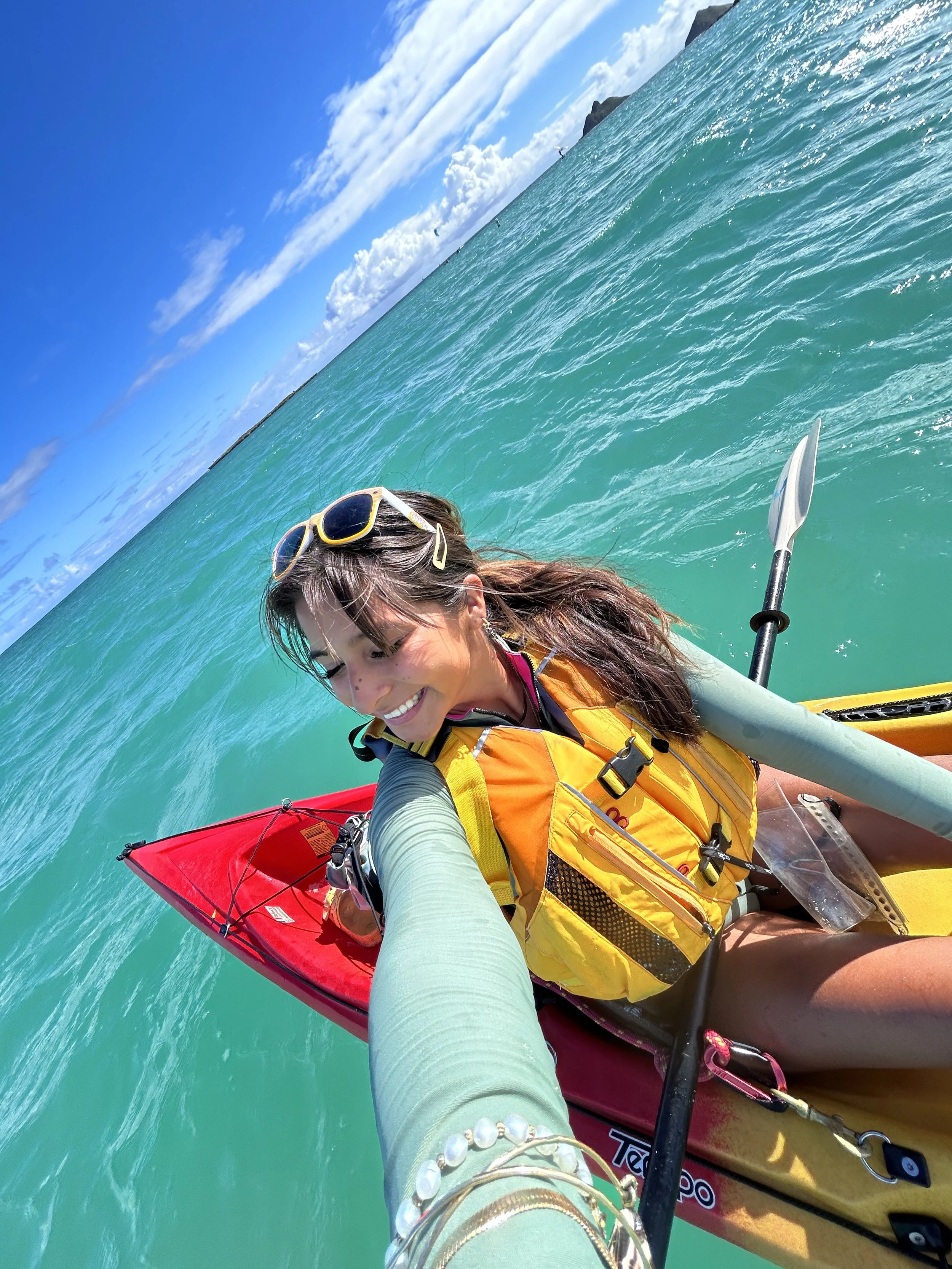 A woman smiling and kayaking on clear blue ocean water with a bright sky and ocean horizon in the background.