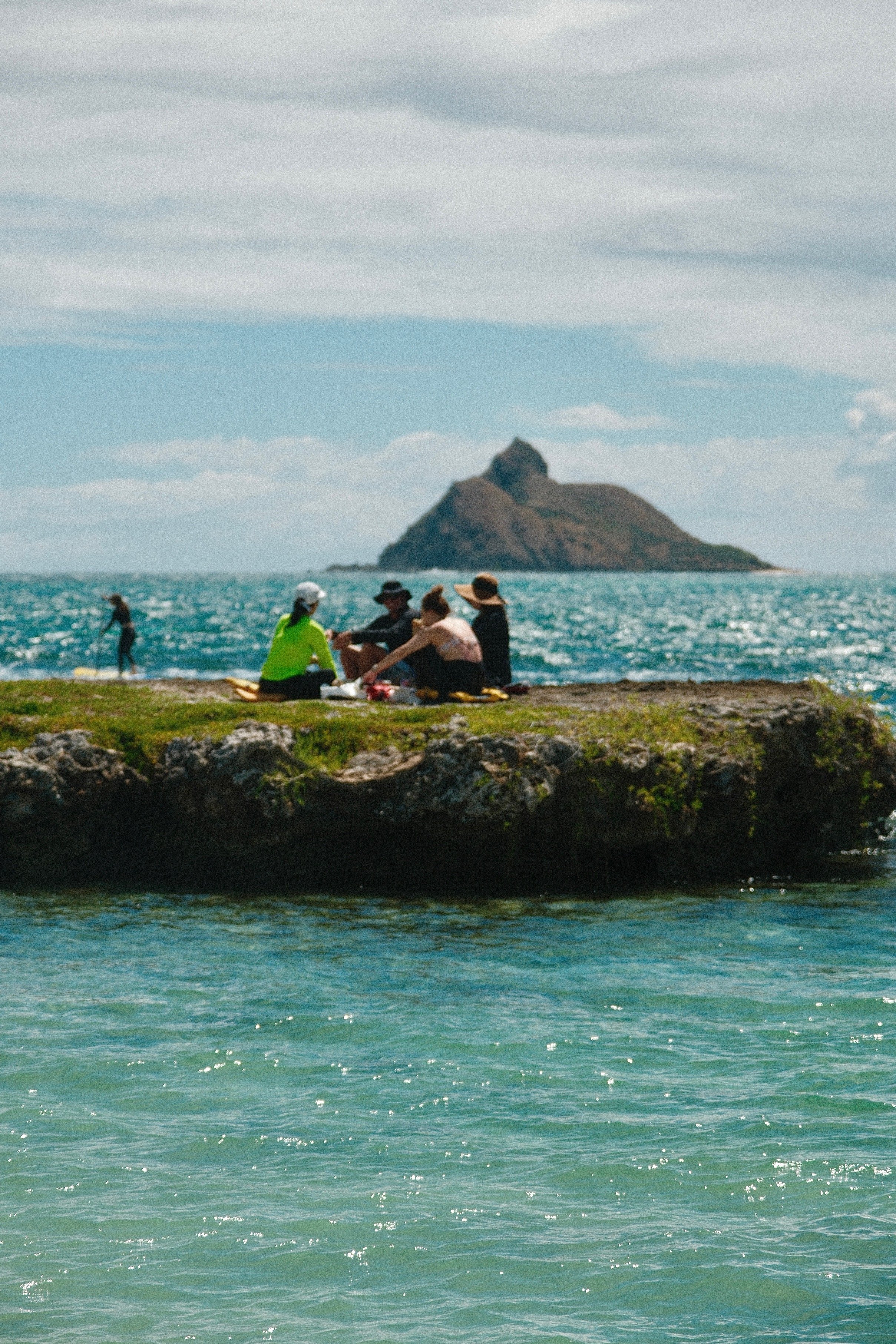People sitting on a grassy rock formation at the beach, with one person in a neon green shirt and others in hats, ocean and an island in the background.