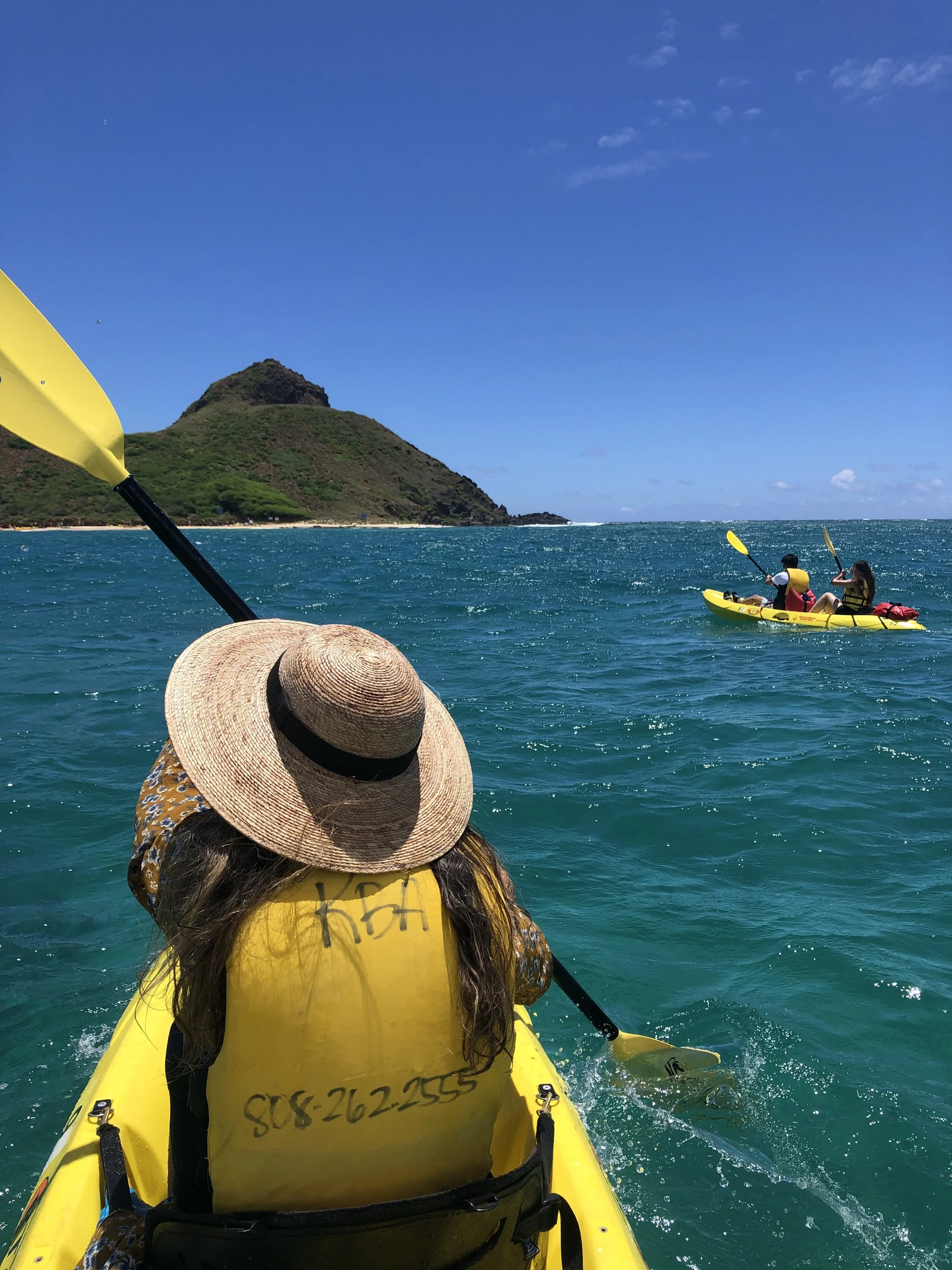 People kayaking on the ocean near a green mountain with a clear blue sky.