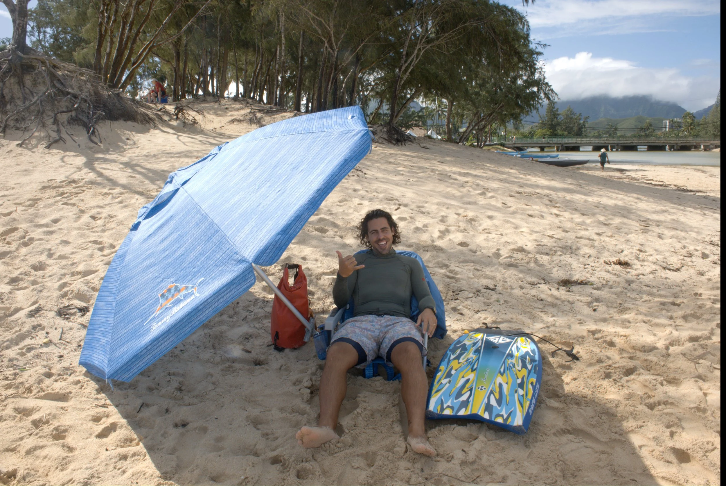 A man is sitting on a beach chair under a blue beach umbrella, smiling and making a 'hang loose' gesture. There is a red bag behind him, and a colorful surfboard is on the sand in front of him. The beach has sand, trees, and water in the background.