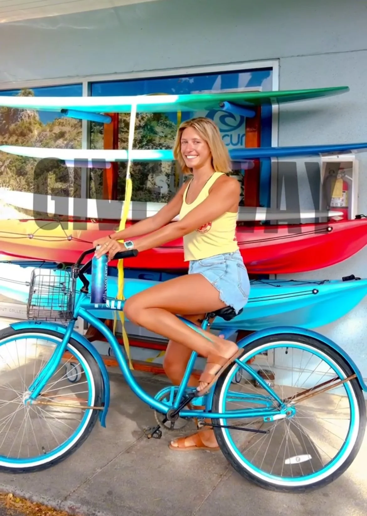 A young woman wearing a yellow tank top and denim shorts rides a blue bicycle with a basket. She is smiling and standing in front of a wall with colorful kayaks and a window.