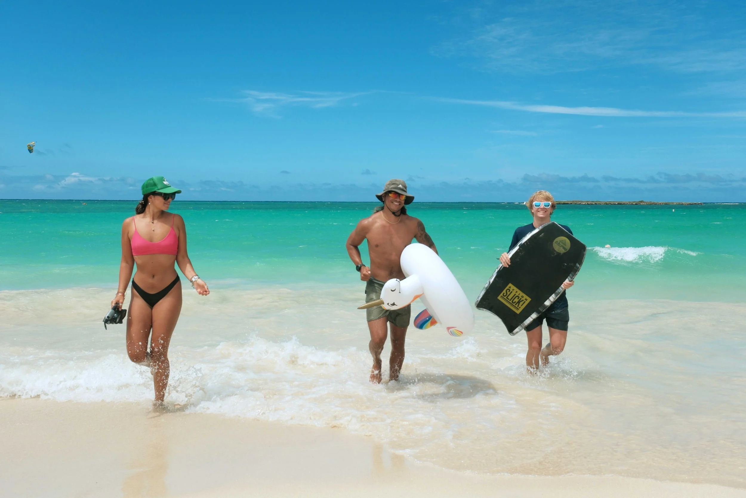 Three people walking out of the ocean at the beach, with two women and one man. The woman on the left wears a pink bikini top, black bikini bottoms, a green cap, and sunglasses. The man in the middle is shirtless, wearing shorts and a sun hat, holding an inflatable unicorn. The woman on the right is wearing a dark rash guard, shorts, and sunglasses, holding a boogie board. The background shows a turquoise sea, blue sky, and a small distant island or landmass.