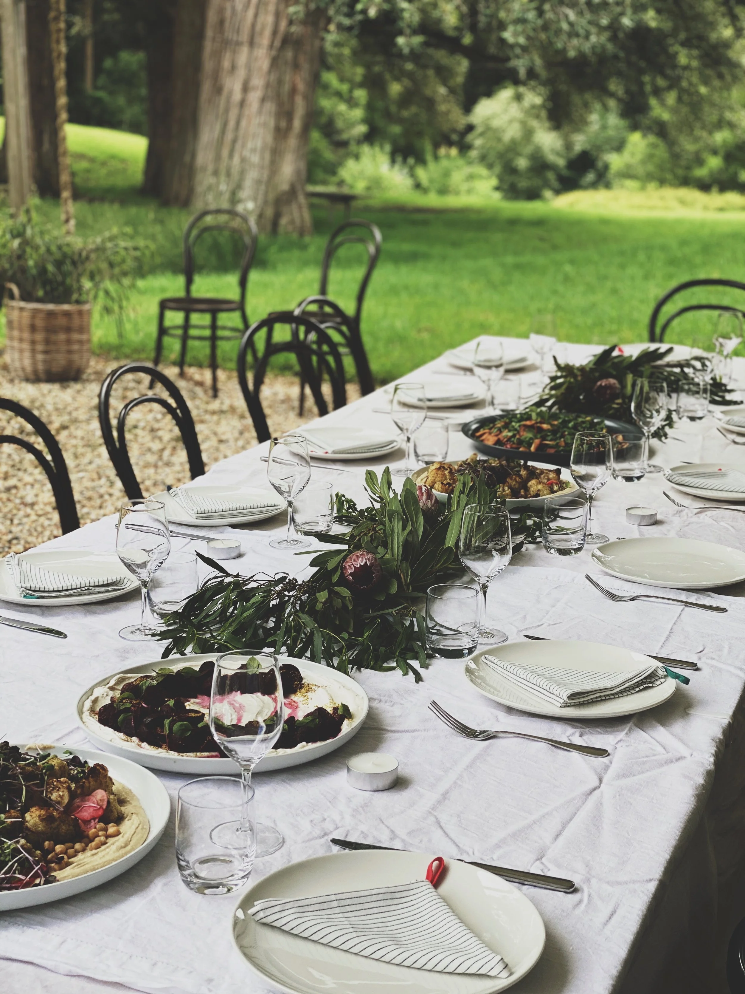 A long outdoor dining table set with plates, glasses, and cutlery, decorated with a greenery centerpiece, surrounded by black chairs, with a grassy area and large trees in the background.