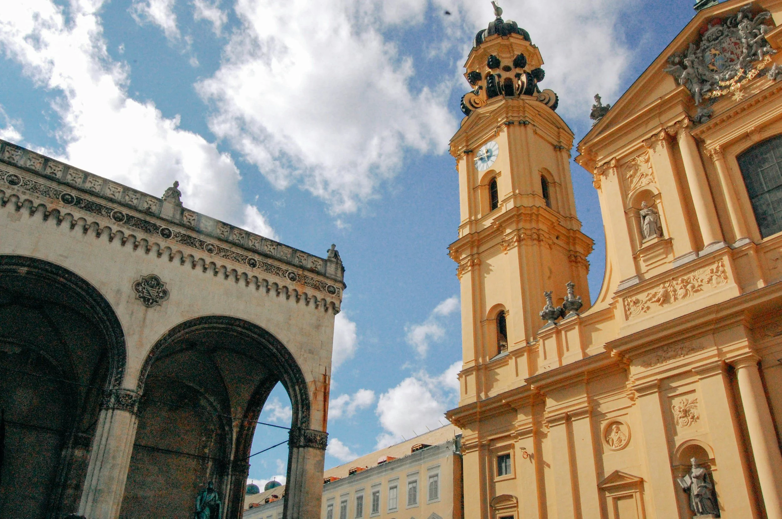 View of a yellow baroque church with a tall clock tower against a blue sky with clouds, and an adjacent structure with arched walkways.