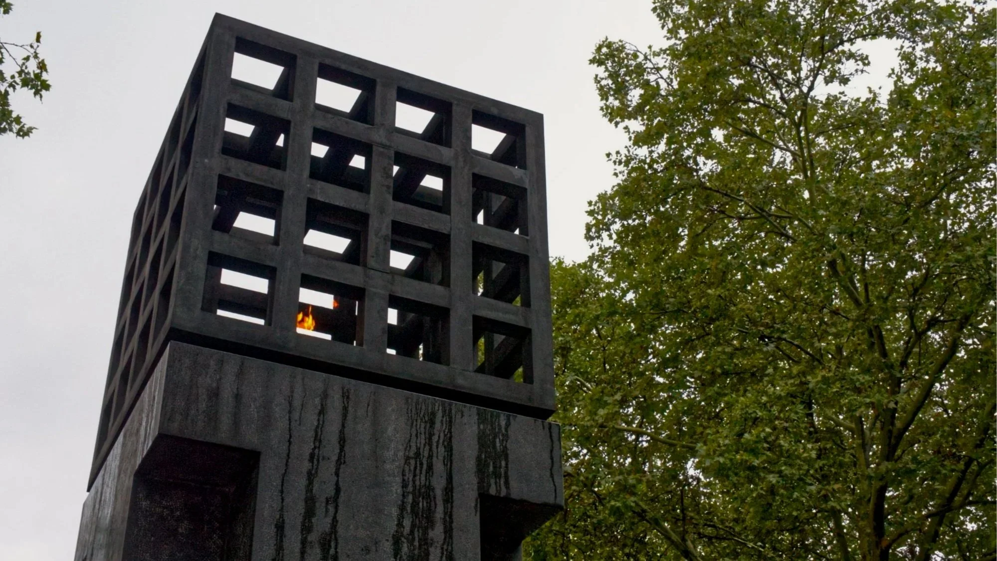 A modern black metal fire pit structure with a back opening, situated outdoors near green trees, with a small visible fire inside.