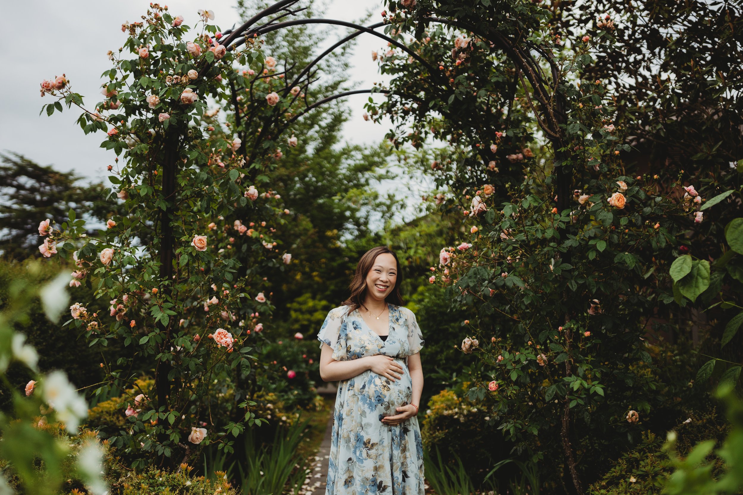 Expecting mother standing beneath a rose-covered arbor during a maternity photo session at the Center for Urban Horticulture in Seattle