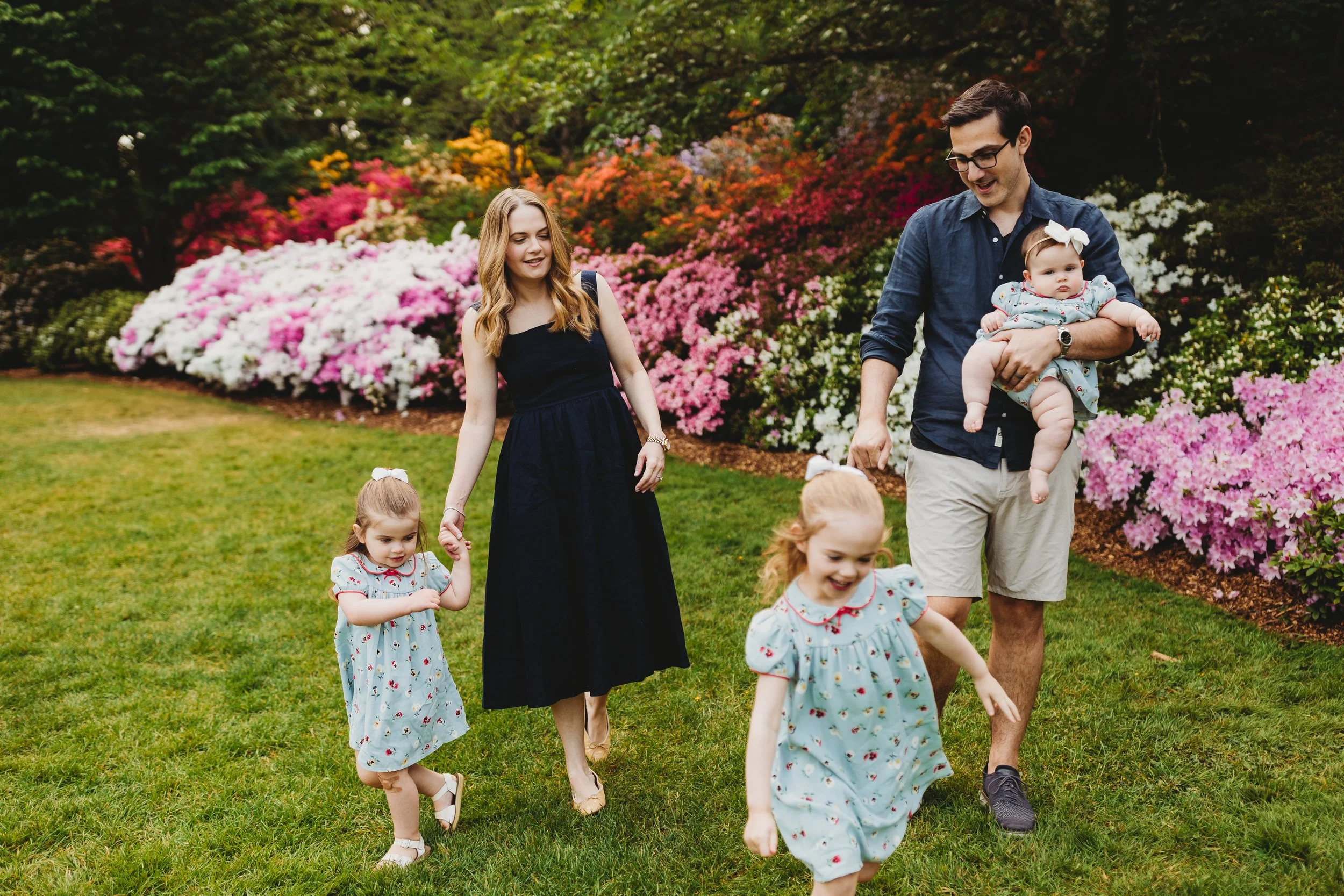 Young family of five — mom in black dress, dad carrying infant, two toddler daughters in matching blue dresses — strolling through a vibrant azalea garden during a spring Seattle family photo session