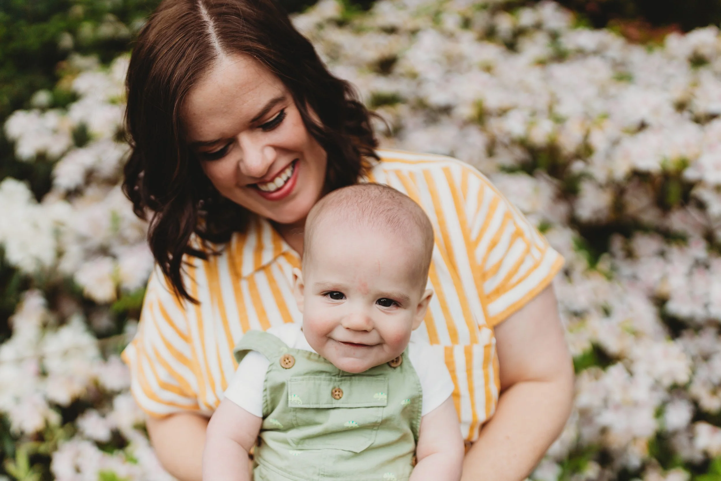 Mom in yellow striped top laughing down at smiling baby in green overalls during outdoor spring mini session in Seattle — white blooms in background