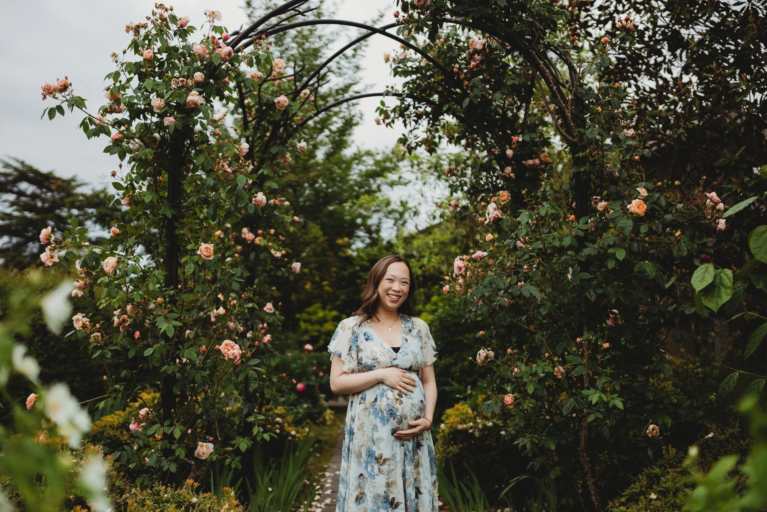Smiling pregnant woman in a light blue floral dress cradling her baby bump while standing beneath a blooming pink rose arch at the Center for Urban Horticulture during a Seattle maternity photography session