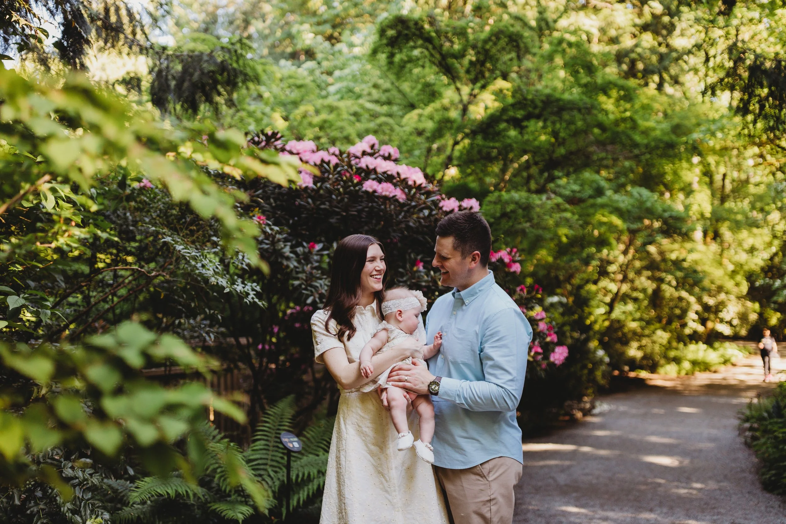 Young couple laughing together while cradling their newborn baby on a sun-dappled garden path lined with pink rhododendrons during a Seattle newborn and family outdoor photography session