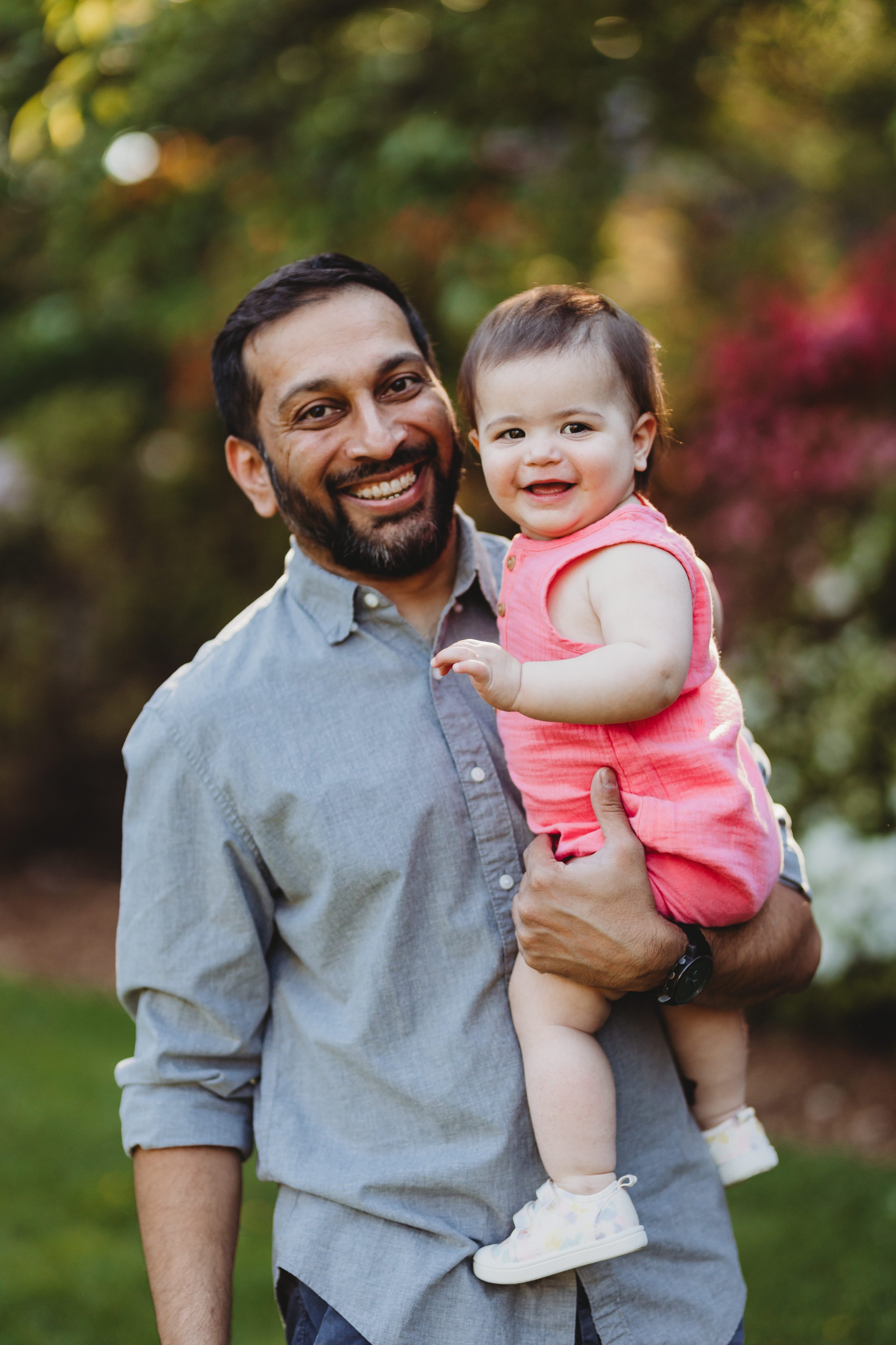 Bearded dad in a grey button-down shirt beaming while holding his happy baby girl in a pink romper during an outdoor Seattle family photography session in a lush garden setting