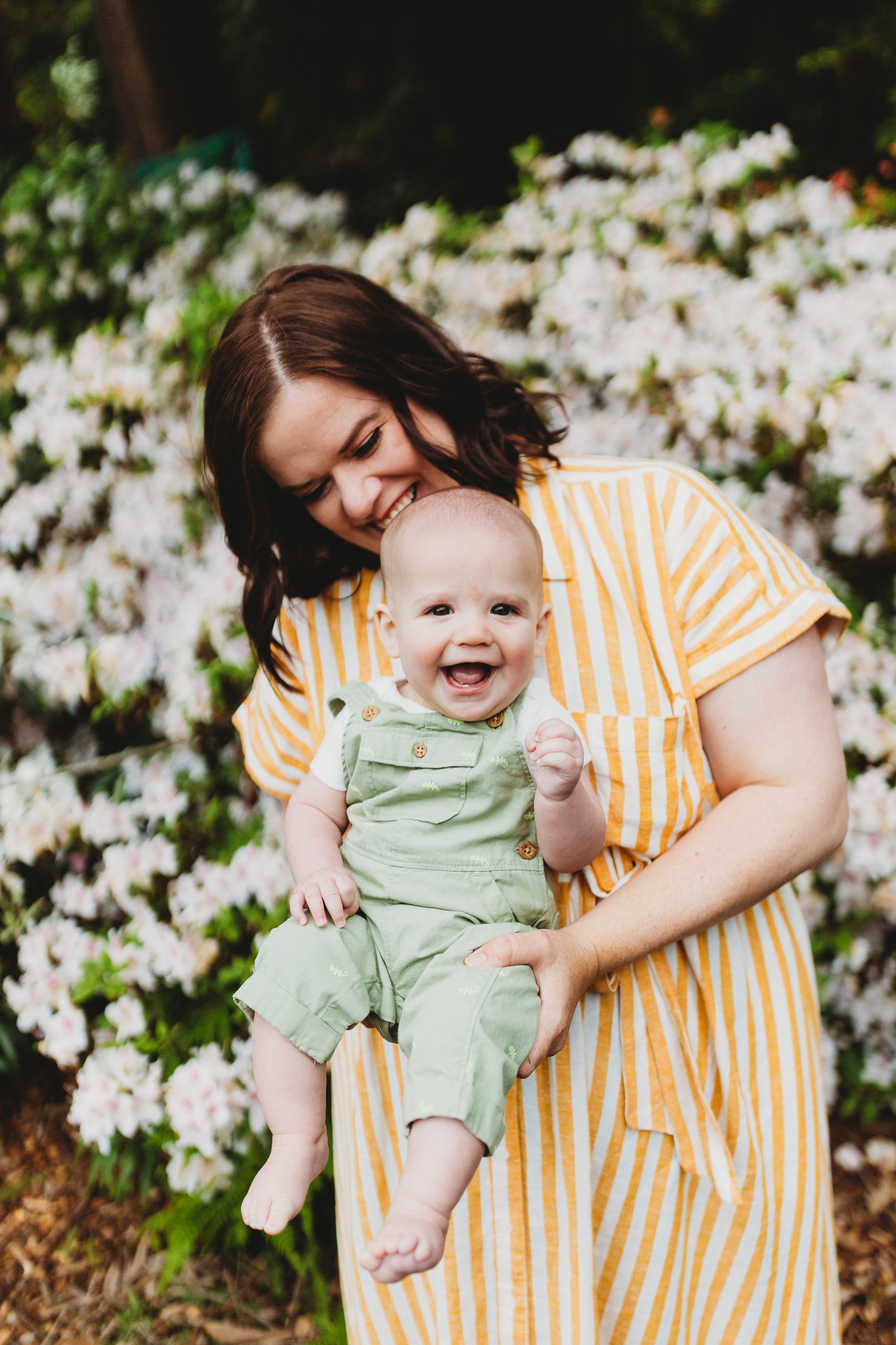 Laughing baby in green overalls held by smiling mom in yellow striped dress during spring mini session at Washington Park Arboretum in Seattle