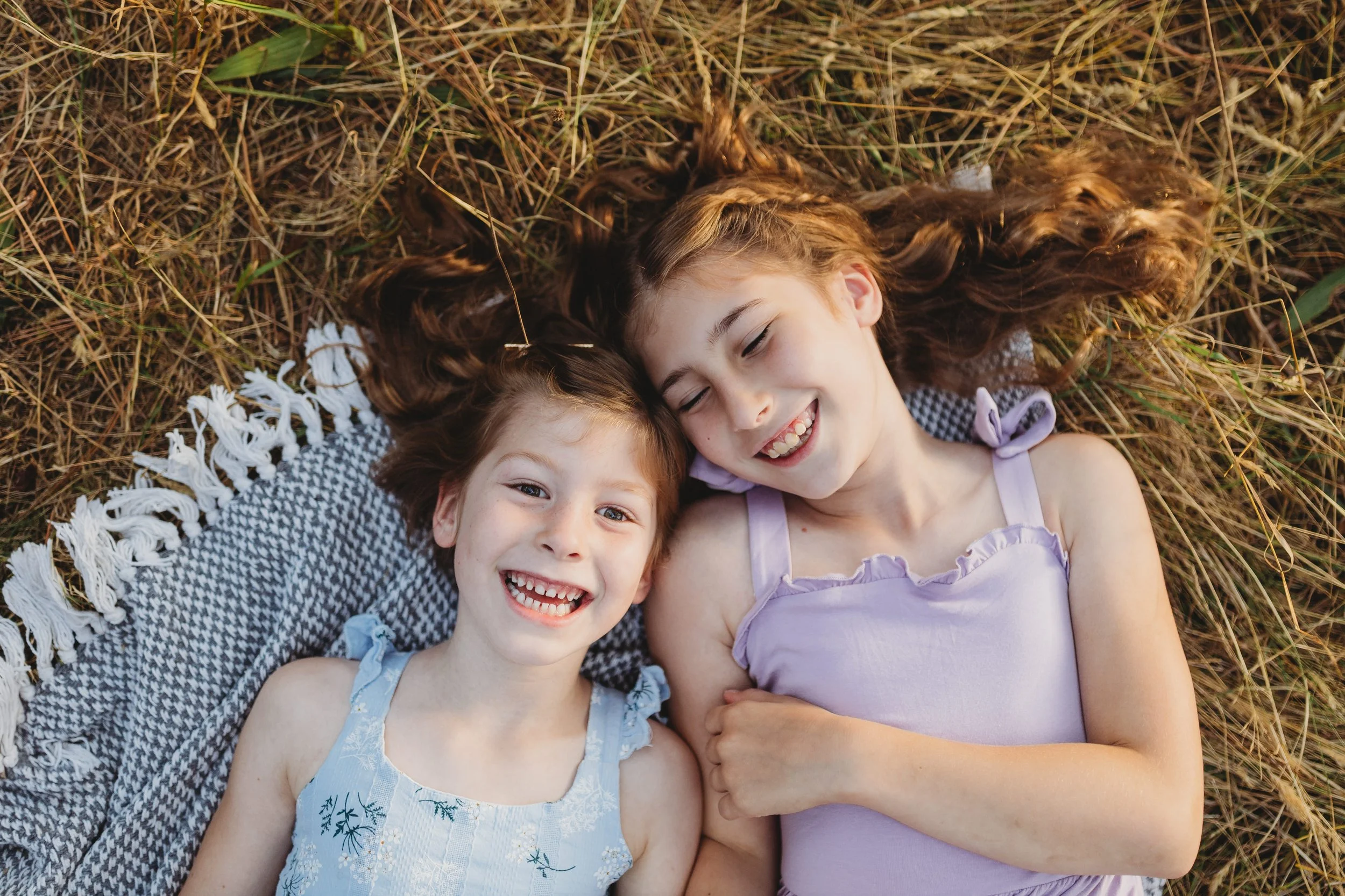 A candid family photo of two sisters laughing together while lying in the grass at Discovery Park in Seattle. This image reflects a relaxed, natural family photography style focused on connection, joy, and real moments.