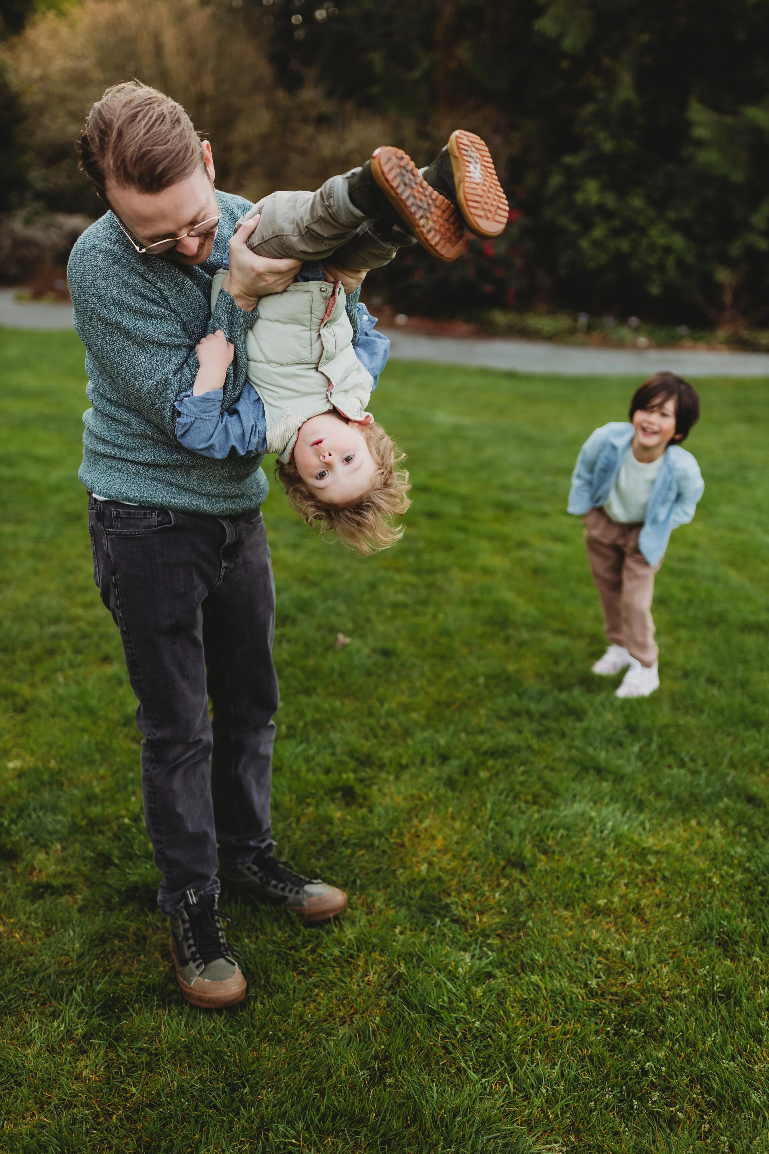 Father laughing while holding his toddler upside down on a green lawn during a candid Seattle family photography session, with an older sibling laughing in the background