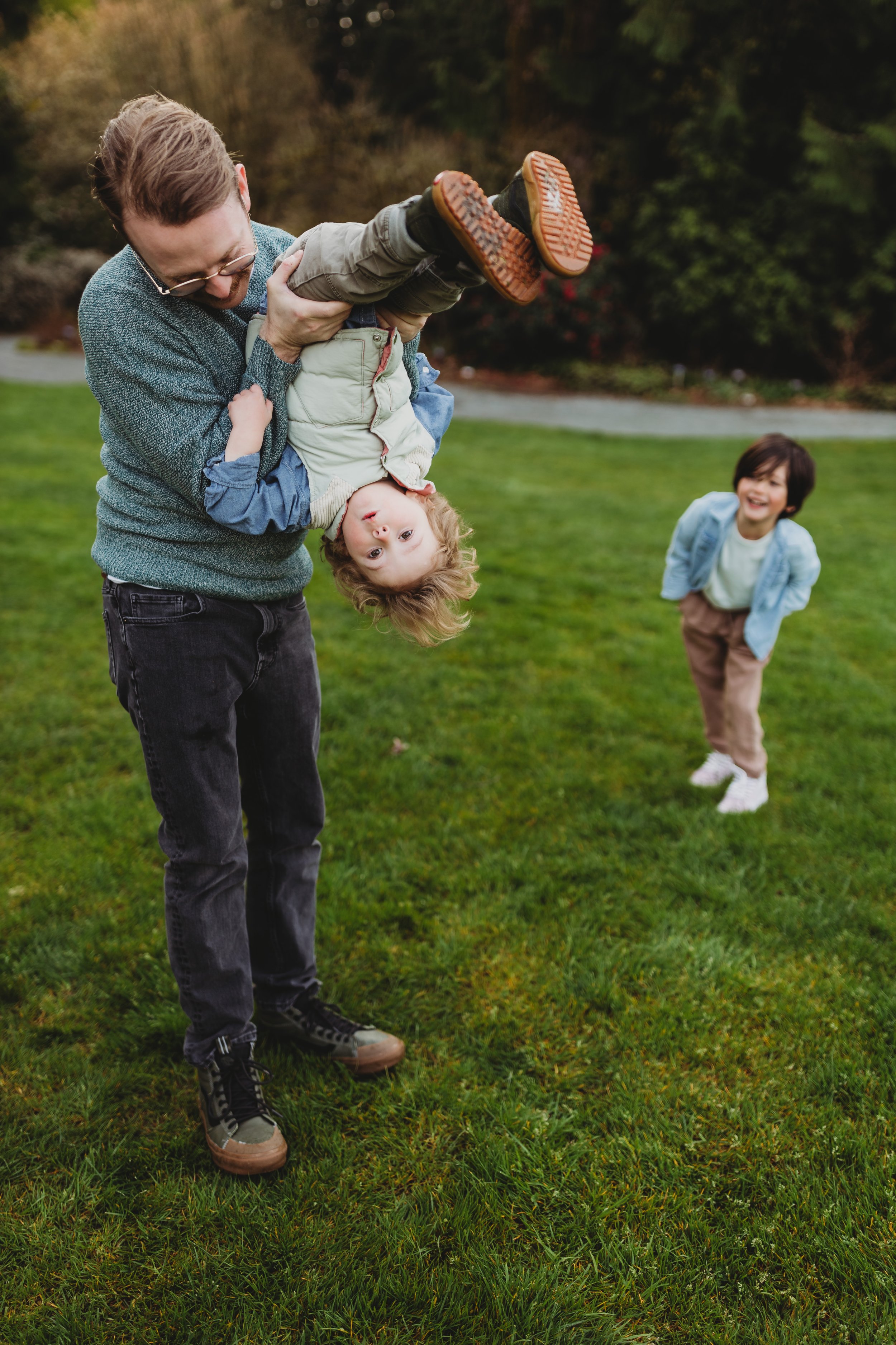 Dad playfully holds toddler upside down while older child laughs in the background during a spring mini session at Bellevue Botanical Garden