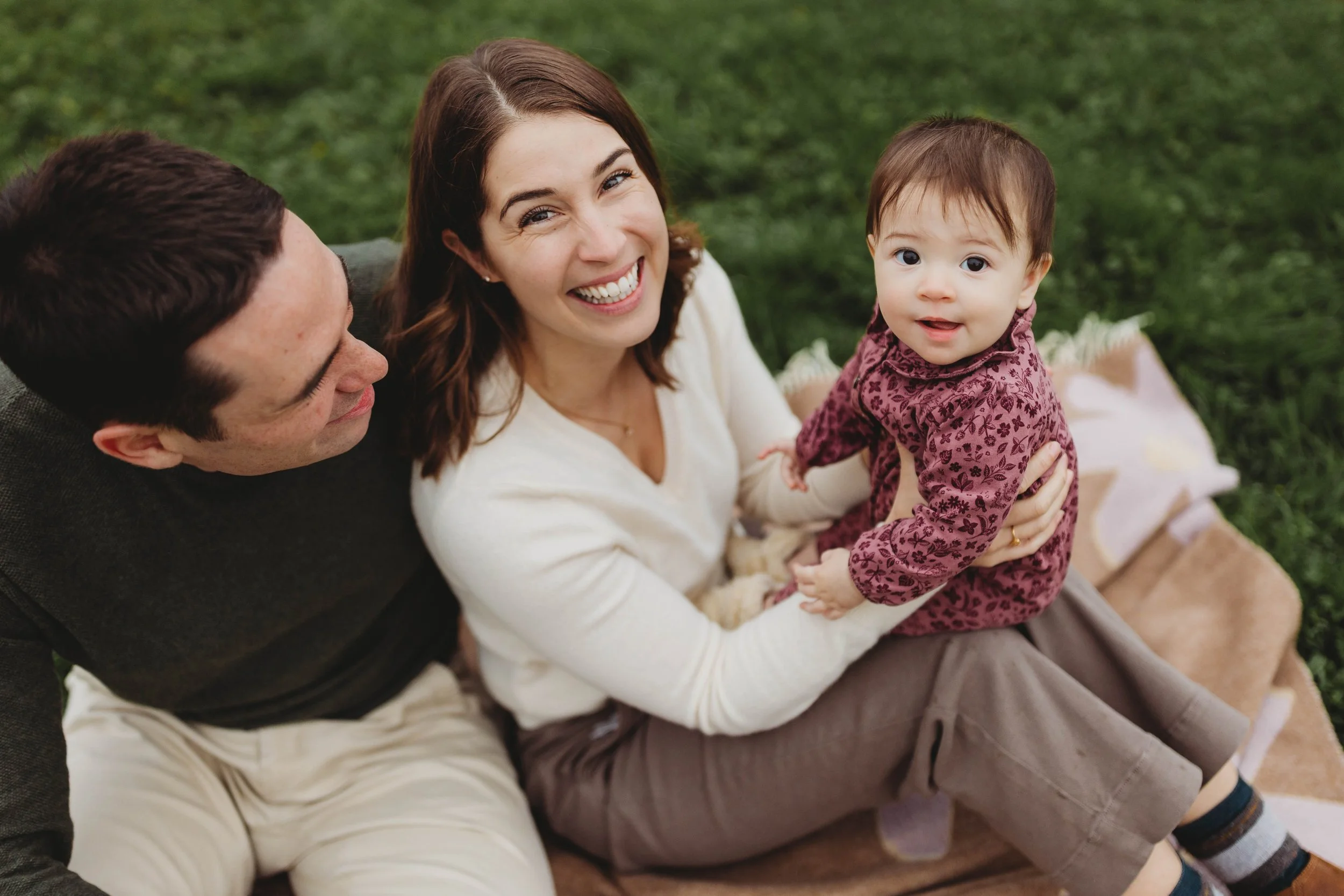 Smiling parents sitting on a blanket holding their baby during an outdoor family photo session at the Center for Urban Horticulture in Seattle