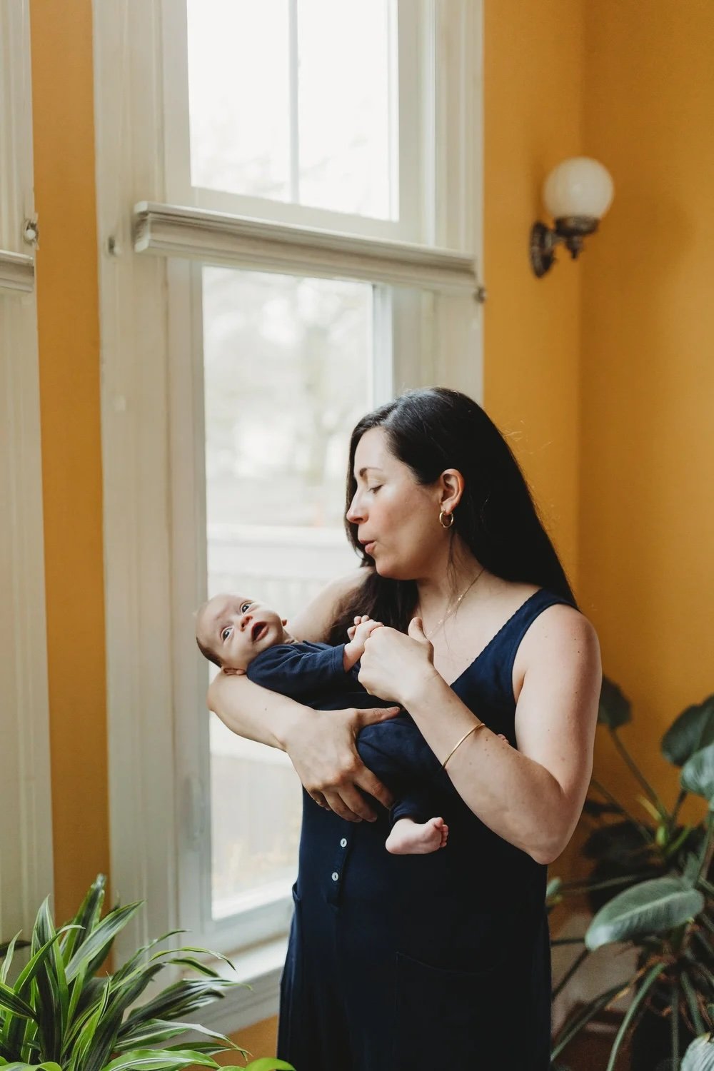 Birthing parent holding newborn by a window during a Seattle in-home lifestyle newborn session, wearing a simple dark dress in natural light.