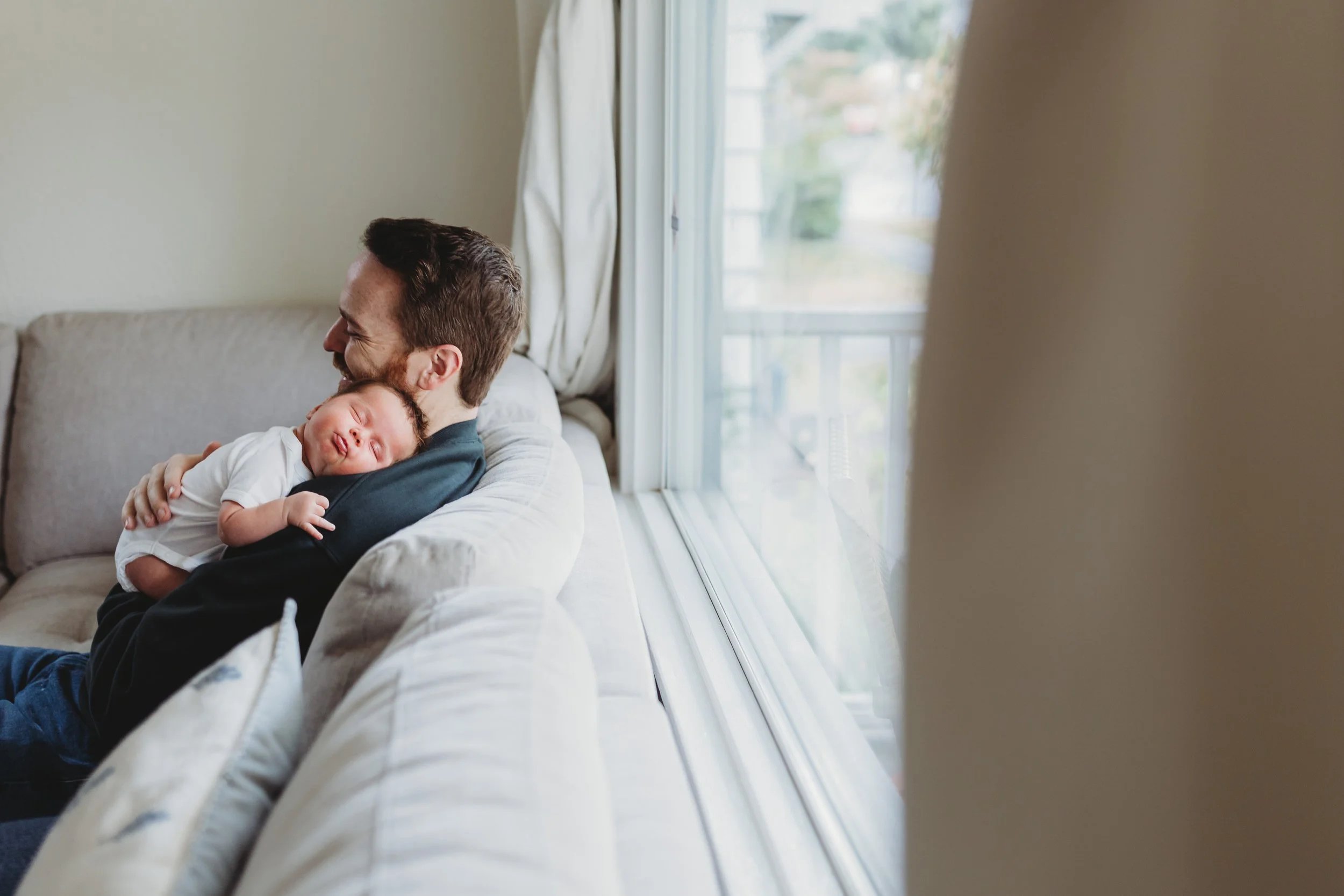 Dad holding sleeping newborn on couch by window during in-home lifestyle newborn photo session in Seattle