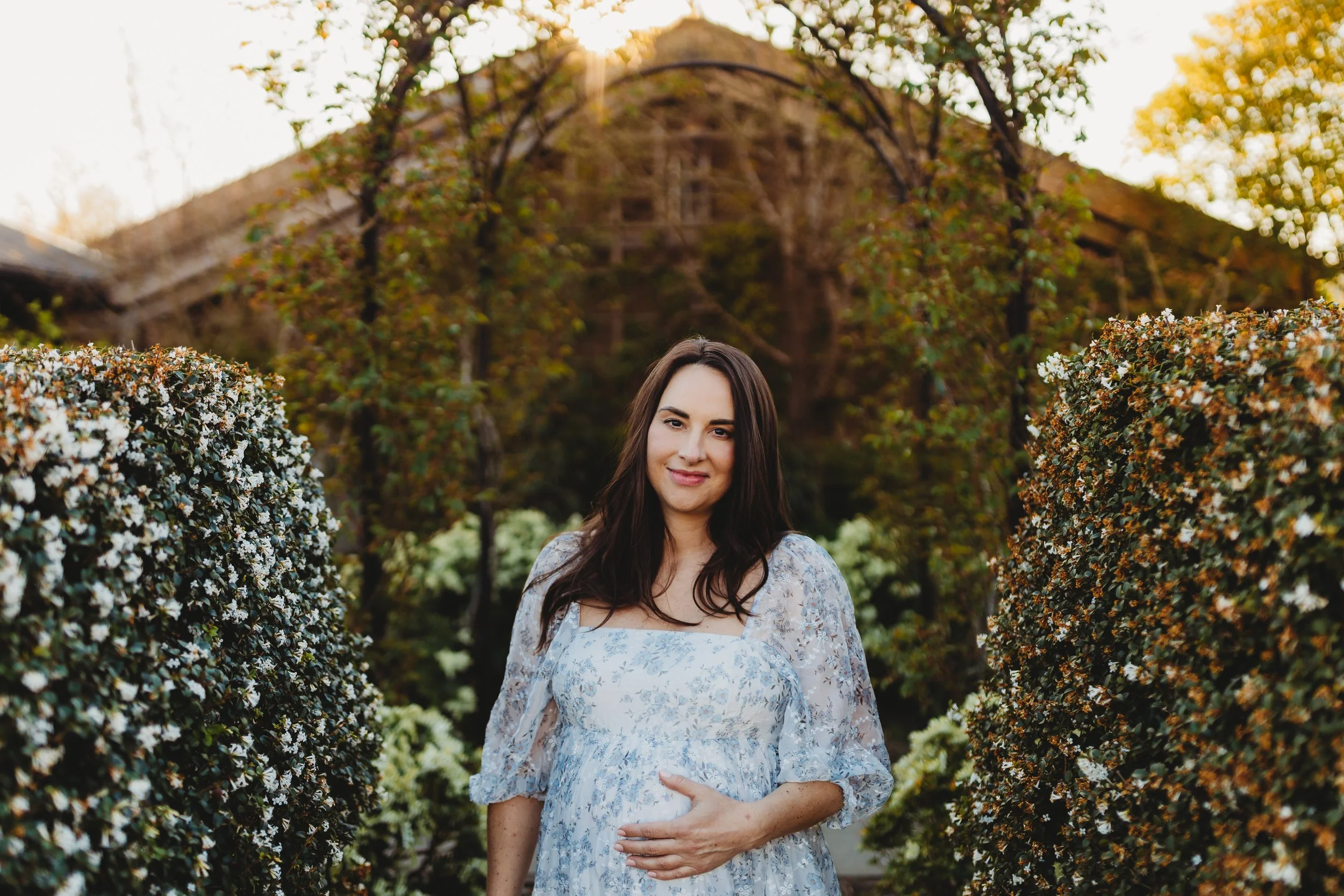 Pregnant woman in a blue floral maxi dress holding her bump under a garden arch at the Center for Urban Horticulture — Seattle maternity photographer