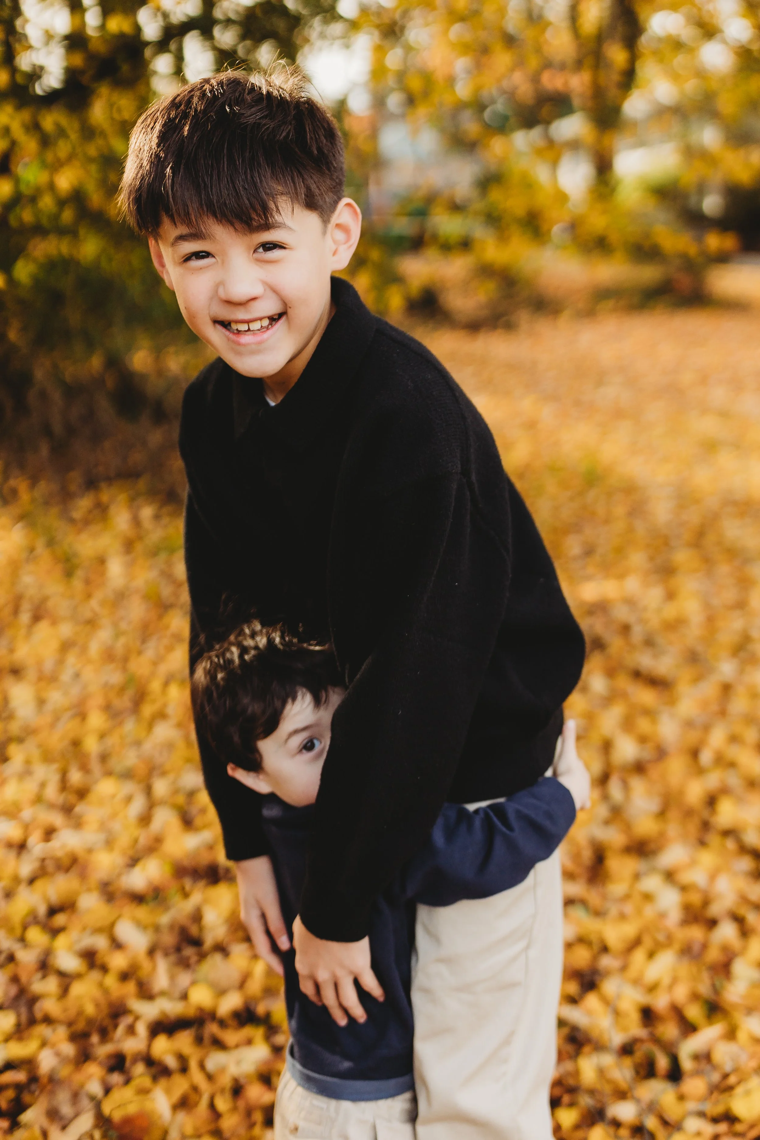 Older brother laughing while his younger brother peeks out from behind him, Ravenna Park fall mini session, Seattle photographer