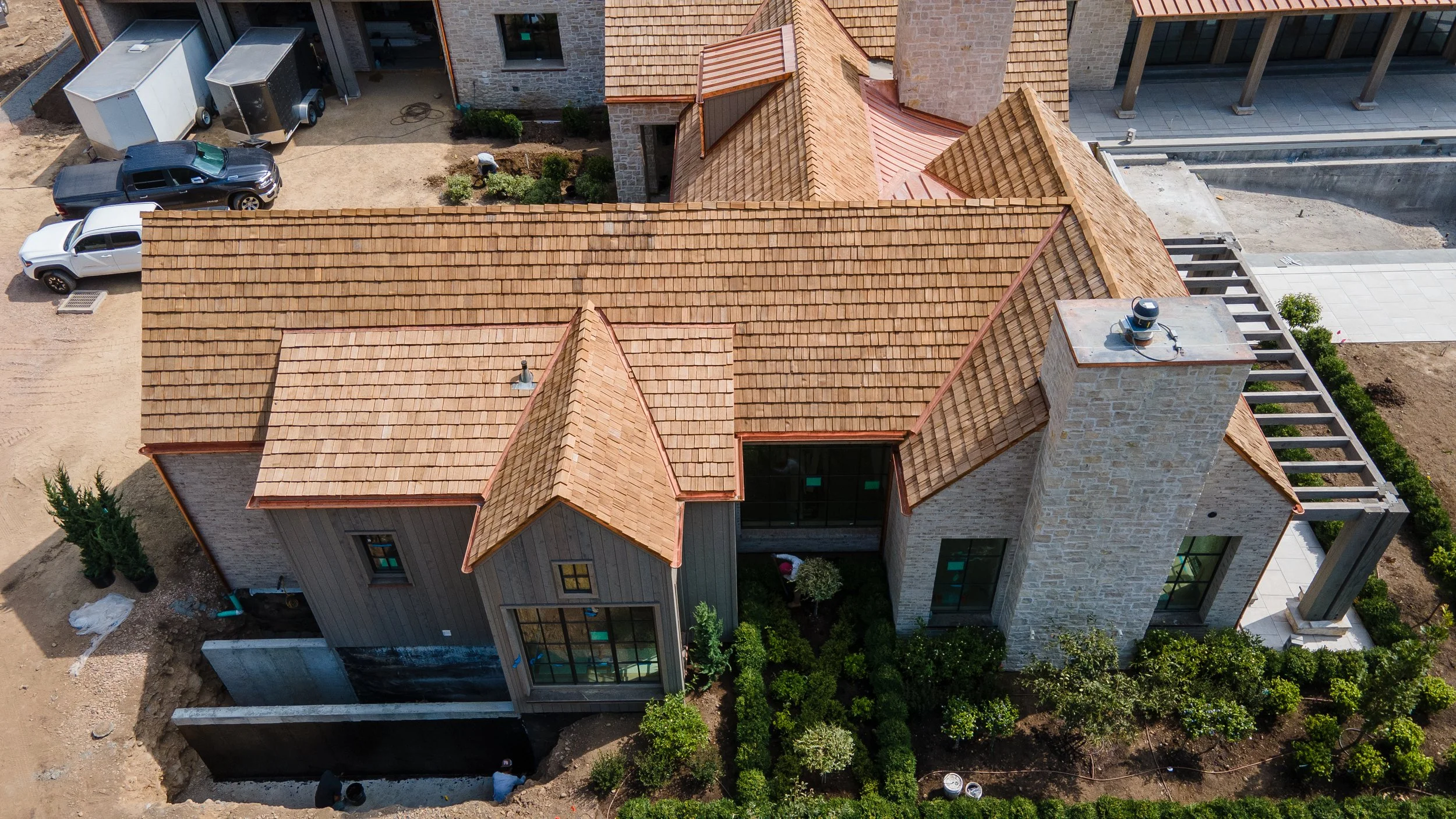 Aerial view of a house under construction with a brown tiled roof, stone chimney, and surrounding green landscaping.