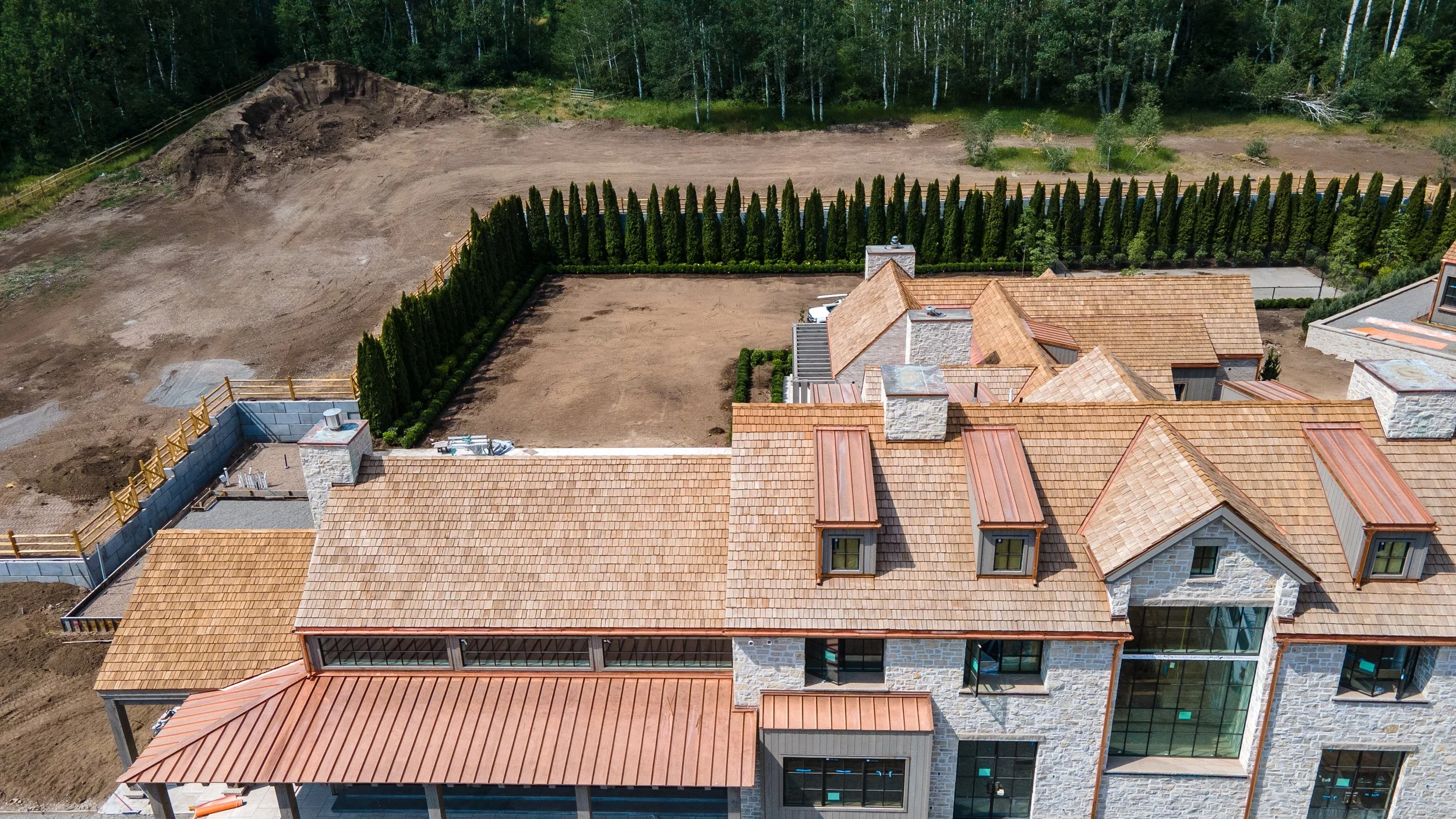 Aerial view of a large house with a red tile roof, surrounded by a landscaped yard with a row of tall, dense evergreen trees and a fenced area. The yard has cleared dirt areas, indicating ongoing landscaping or construction.