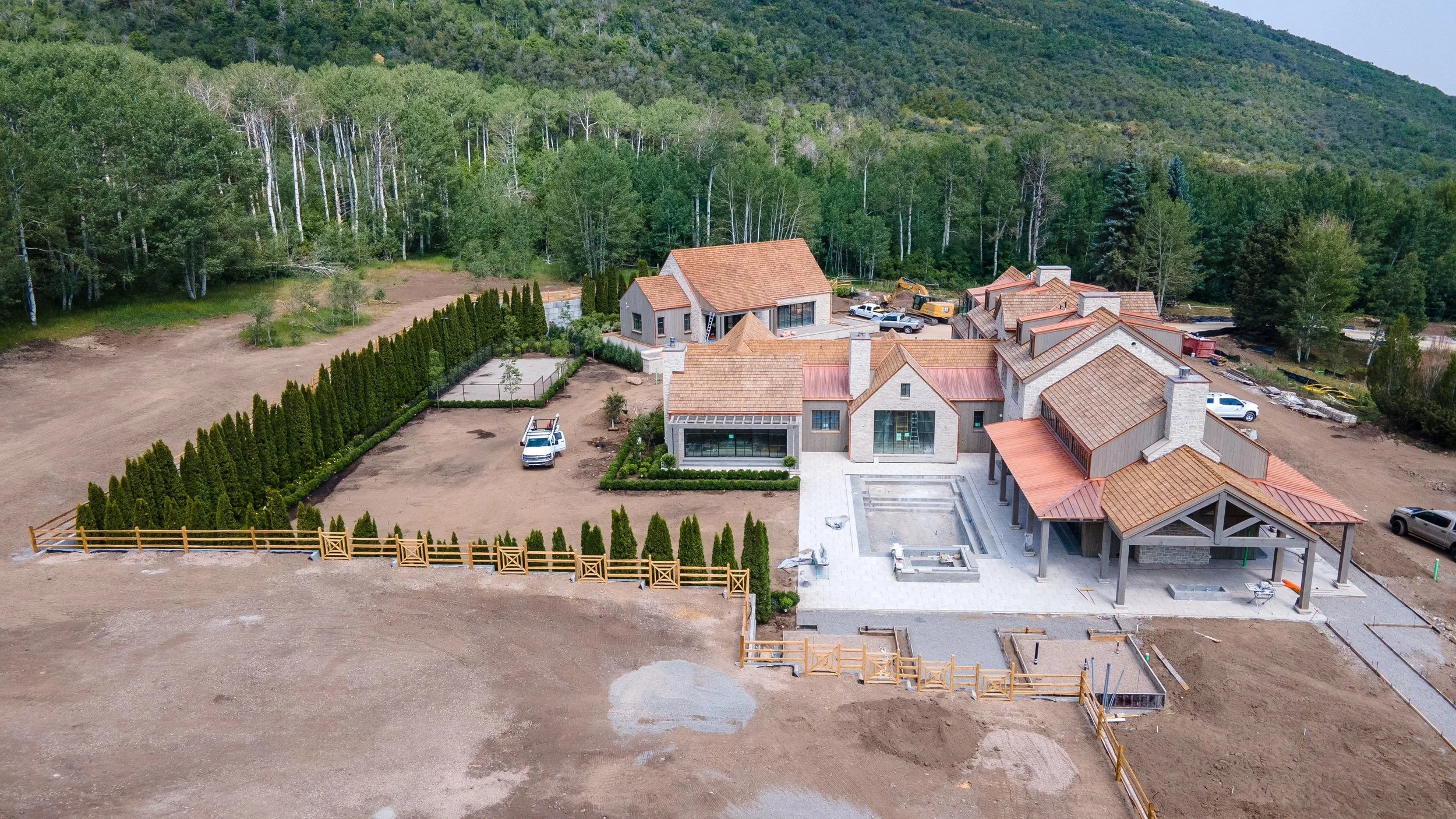 An aerial view of a large residential development under construction surrounded by a wooded area. Several houses with orange-tiled roofs are partially built, with construction vehicles and materials visible. The property features a fenced yard with n
