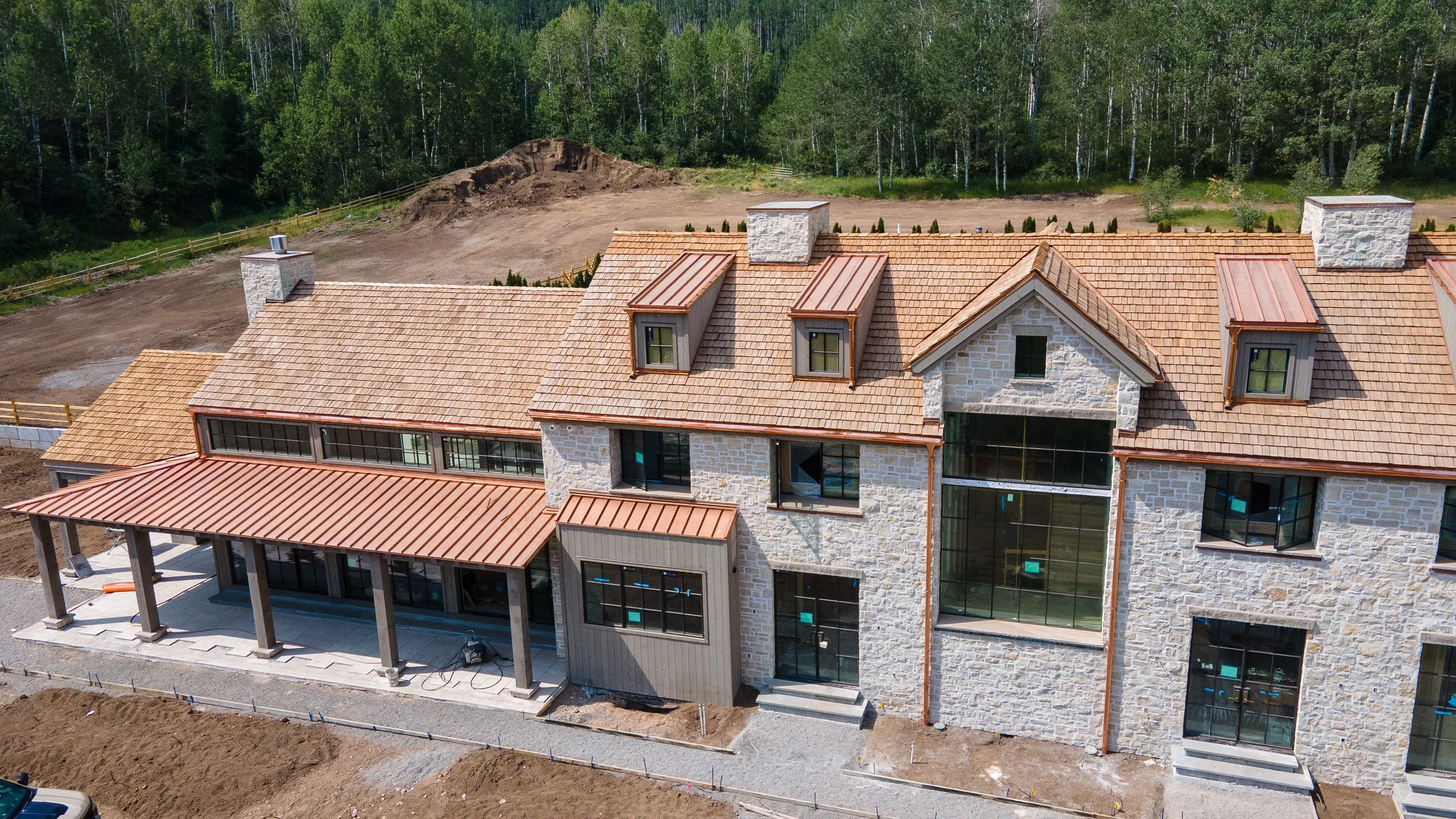 A large residential building under construction with a stone facade, a tiled roof, and multiple dormer windows. The building has a mix of stonework and siding, and is surrounded by dirt and construction materials. In the background, there is a wooded