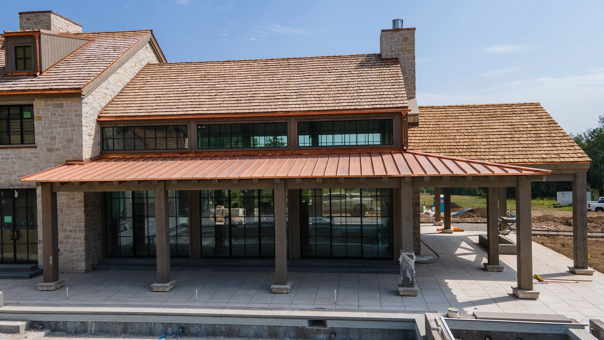 Under construction house with stone walls, large glass doors, a second-story horizontal window, and a rust-colored metal roof, with a patio area under development.