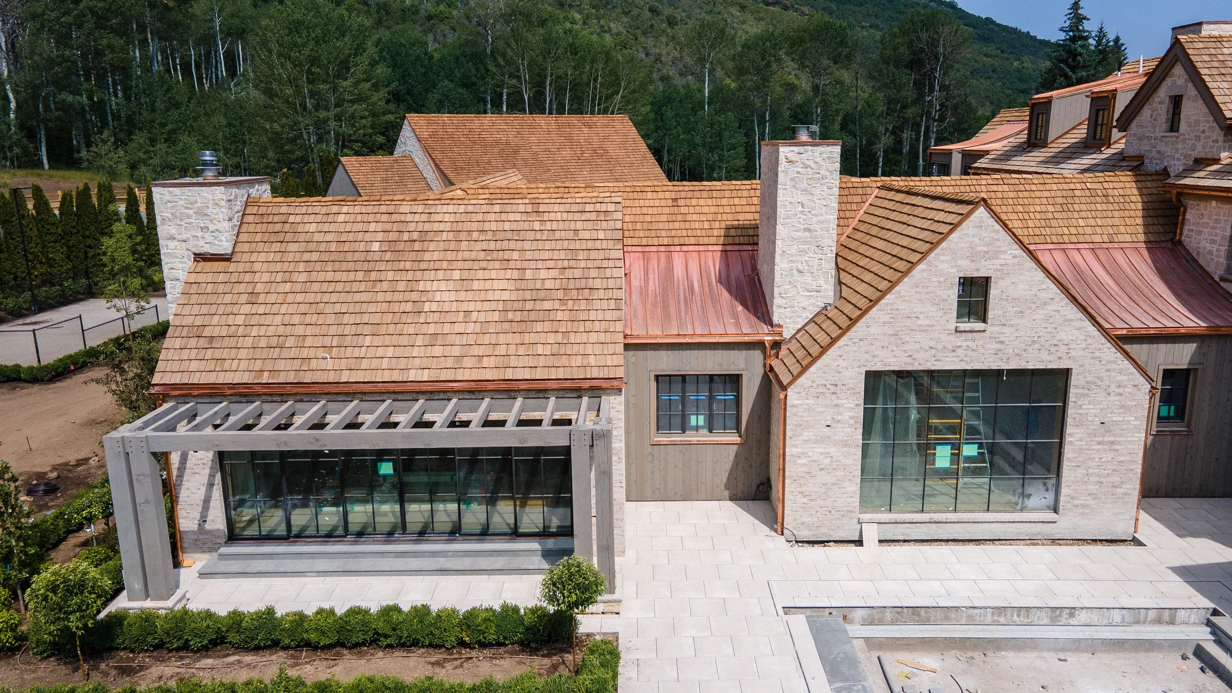 An aerial view of a partially constructed house with a tan tile roof, large windows, and landscaped surroundings, with a backdrop of trees and hills.