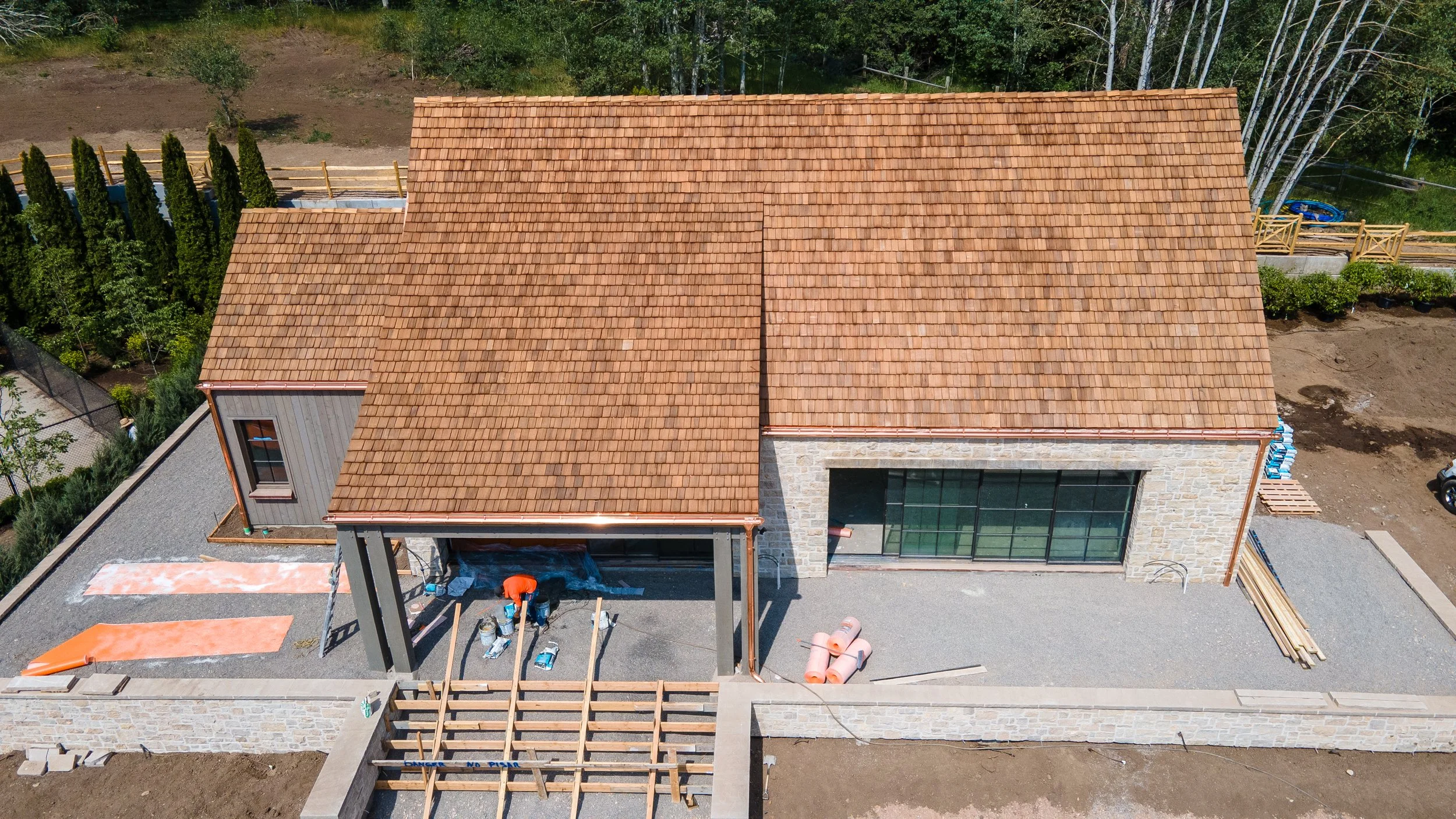 An aerial view of a house under construction with a tiled roof, stone and wood exterior walls, and a spacious gravel-covered backyard. Construction materials and workers are present in the yard.