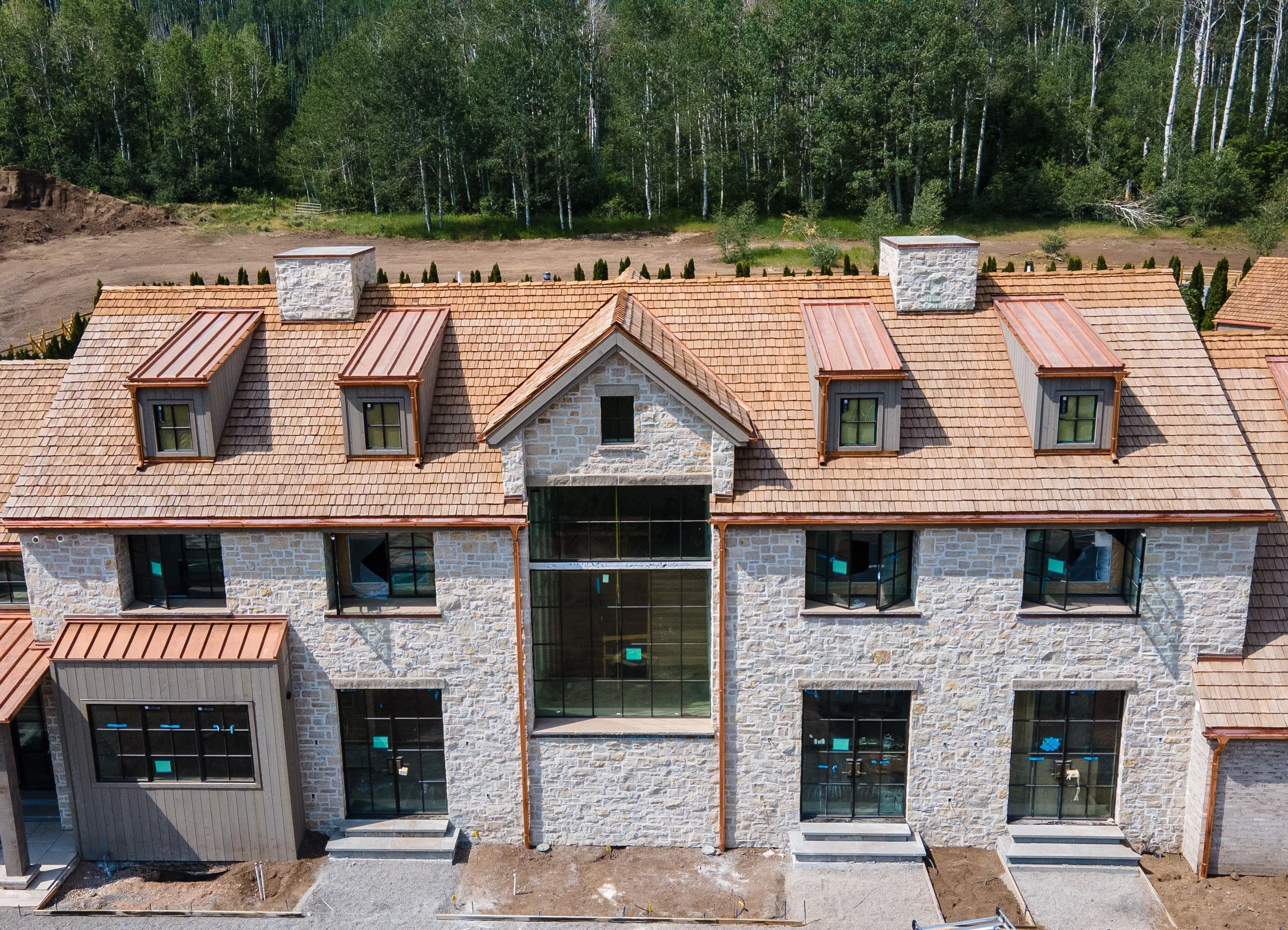 A new multi-story house under construction with a stone exterior and a tiled gable roof, featuring dormer windows and large glass windows, set against a green wooded background.
