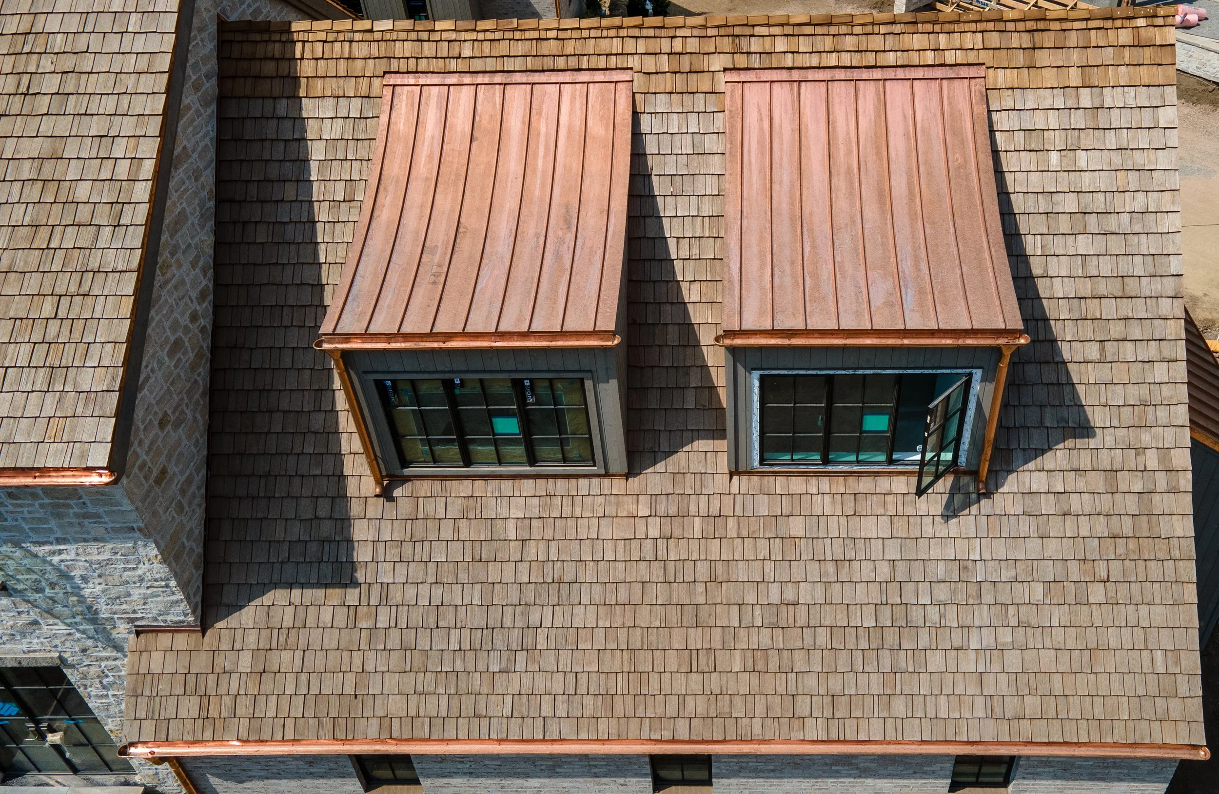 Aerial view of a residential roof with two dormer windows, copper roofing, and varied shingles. Shadows on the roof cast from the sunlight.