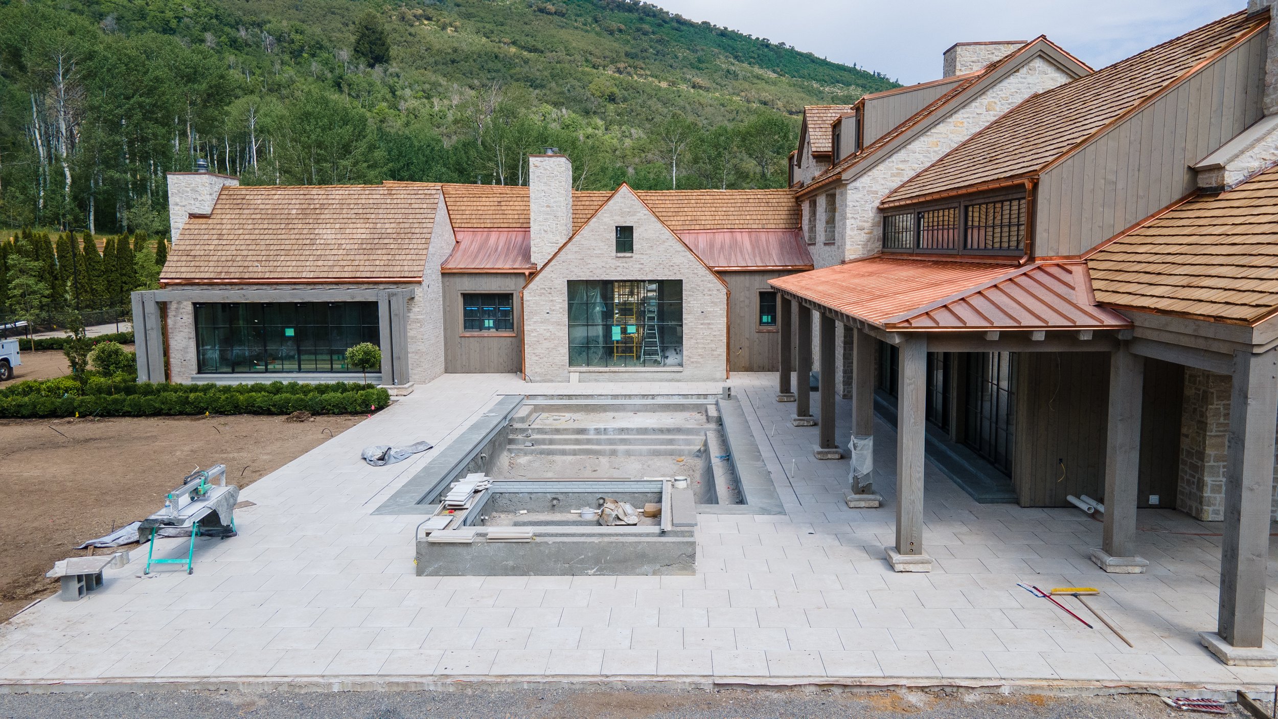 Under construction backyard with a large paved patio, steps leading into a pool area, and a house with brick and wood siding, large windows, and a covered porch, surrounded by greenery and hills in the background.