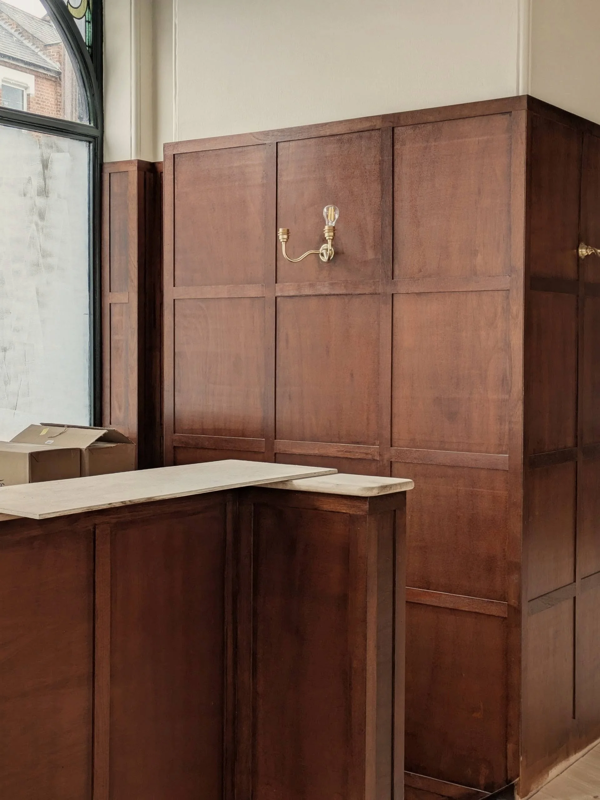 Fleet Tavern interior refurbishment by Polysmiths — heritage timber frame with antique clock as bar backdrop, deep green surfaces, stained dark timber panelling and brass wall lights, Hampstead NW3