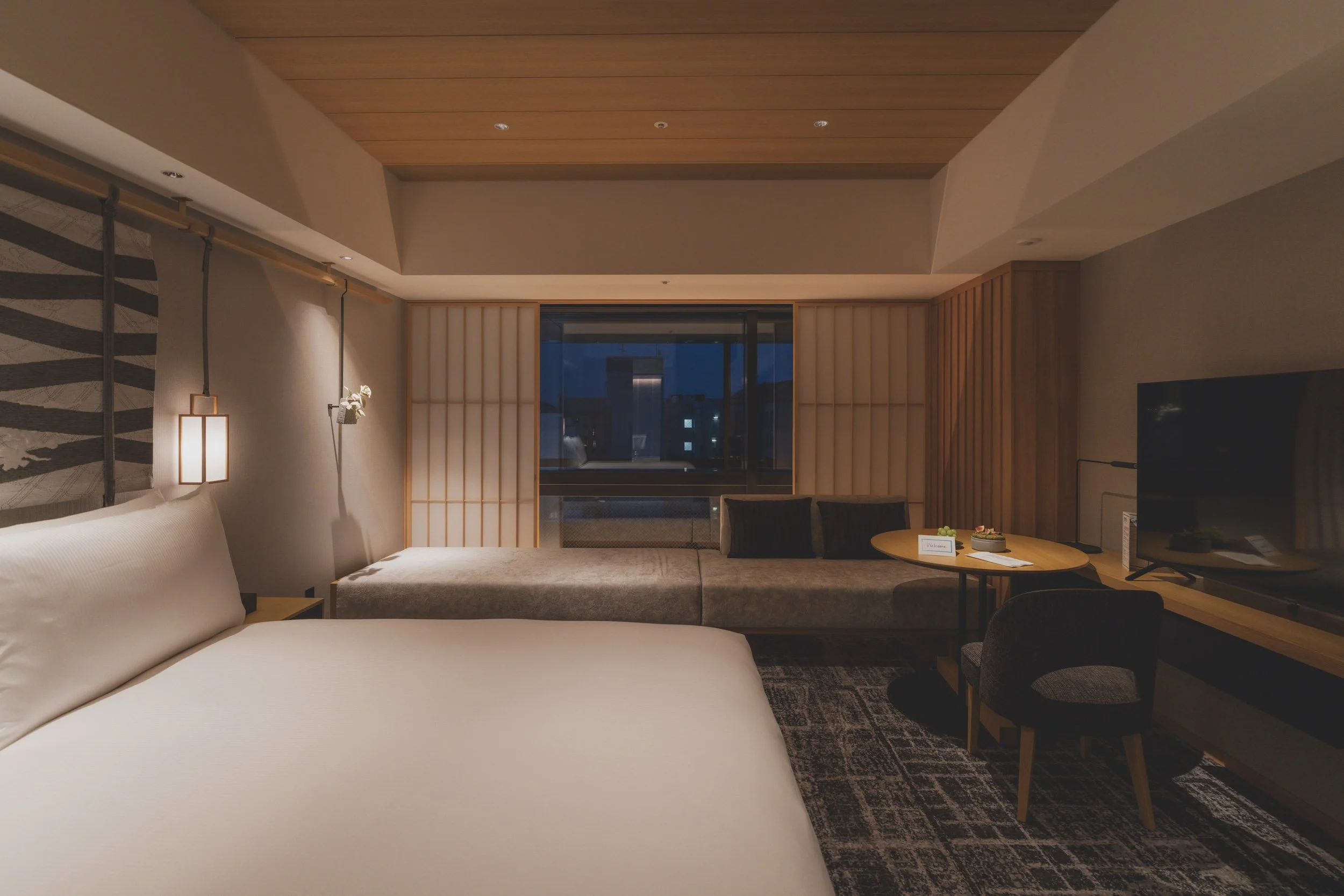 Night view of a contemporary hotel room with a large bed, a sitting area with a window, and a flat-screen TV.