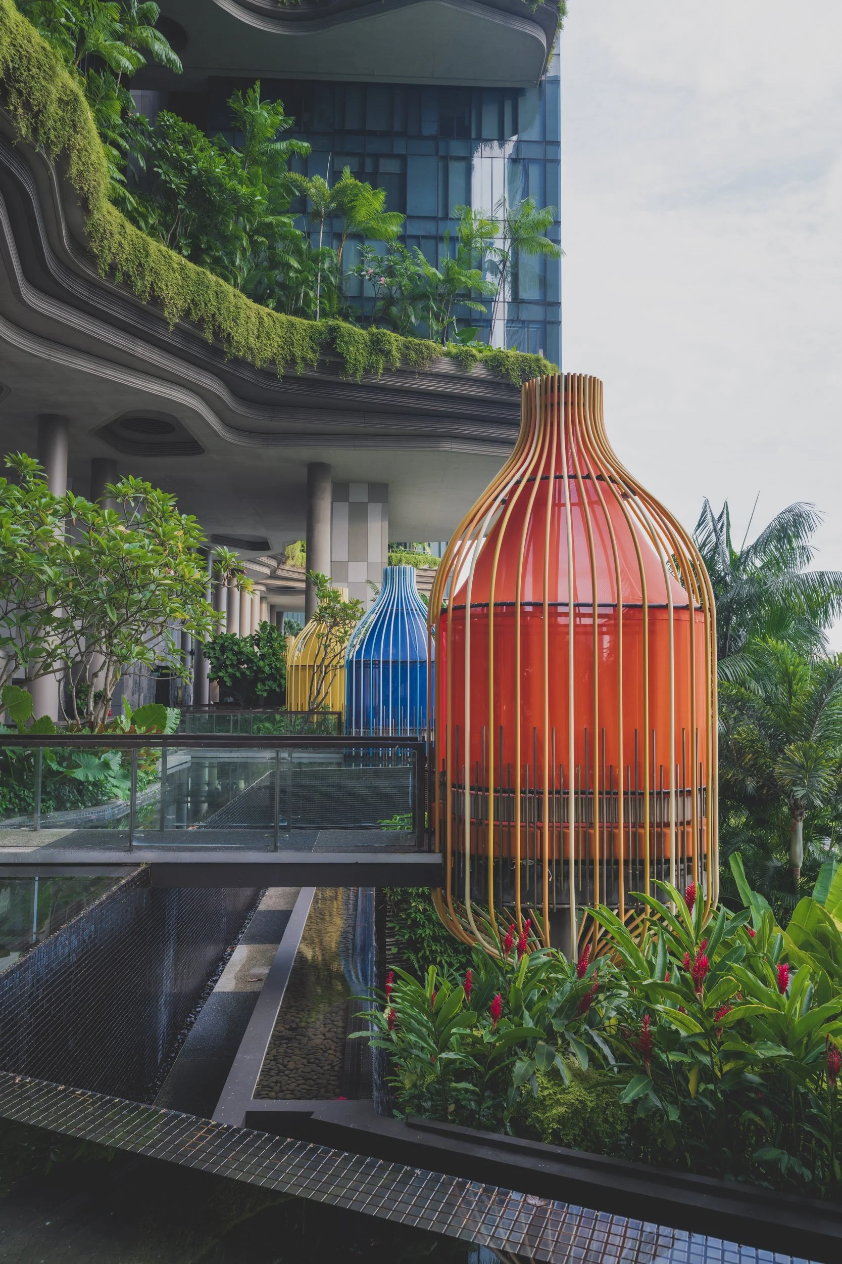 Colorful birdcage-shaped structures in an urban garden with modern buildings in the background.