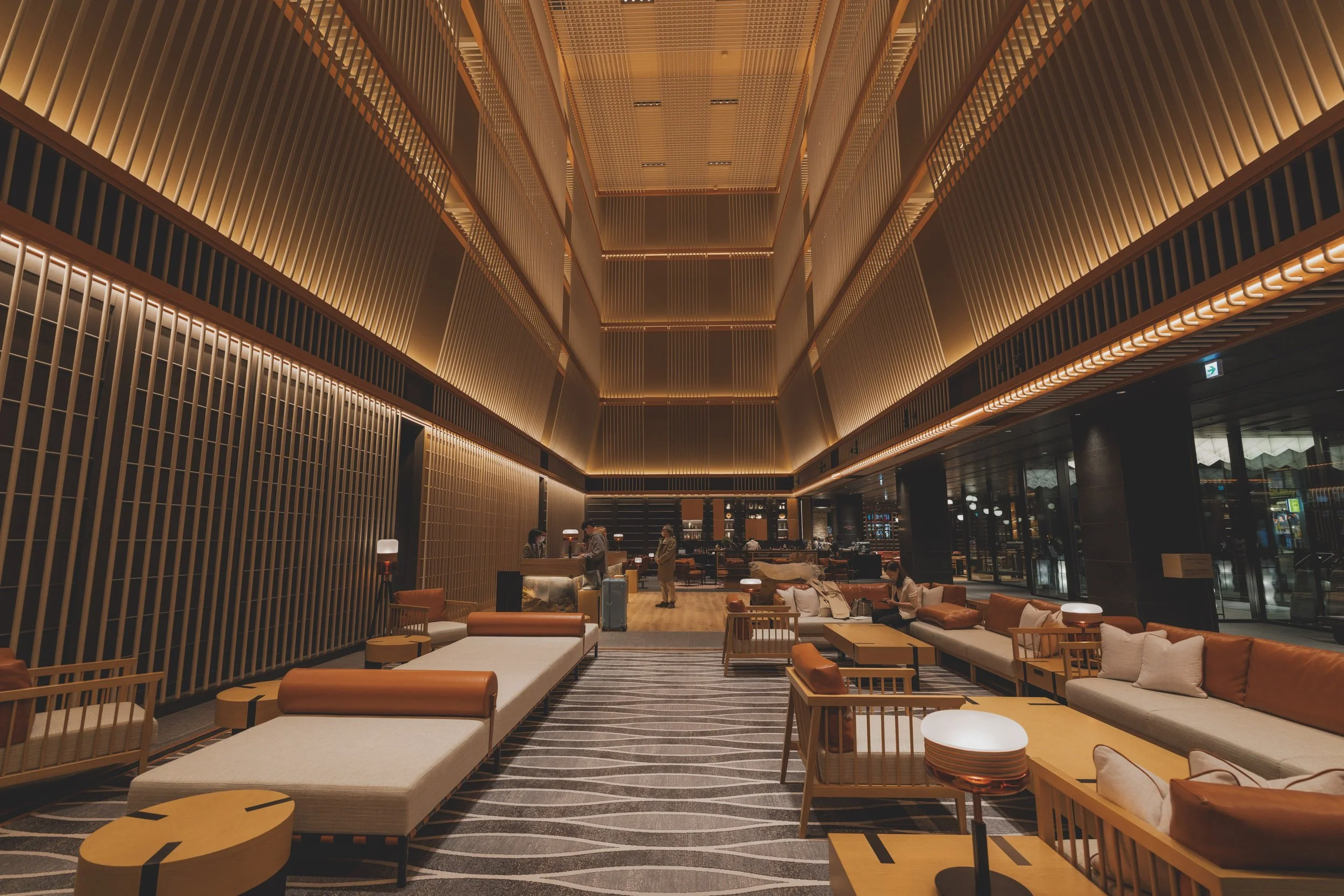 Interior of a modern, upscale hotel lobby with high ceiling, wooden accents, and warm lighting. Lounge area with sofas, chairs, and small tables, with people sitting and walking around.