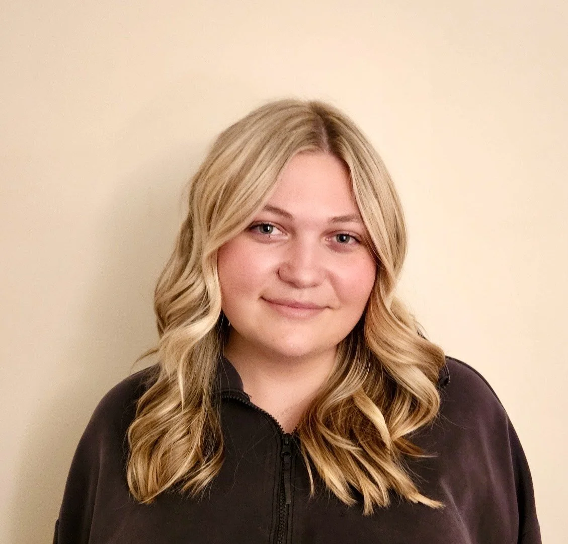 Headshot of Elizabeth Maloney, LMSW, therapist at Upstate Counseling, smiling softly with shoulder-length blonde wavy hair, wearing a dark zip-up top against a light neutral background.