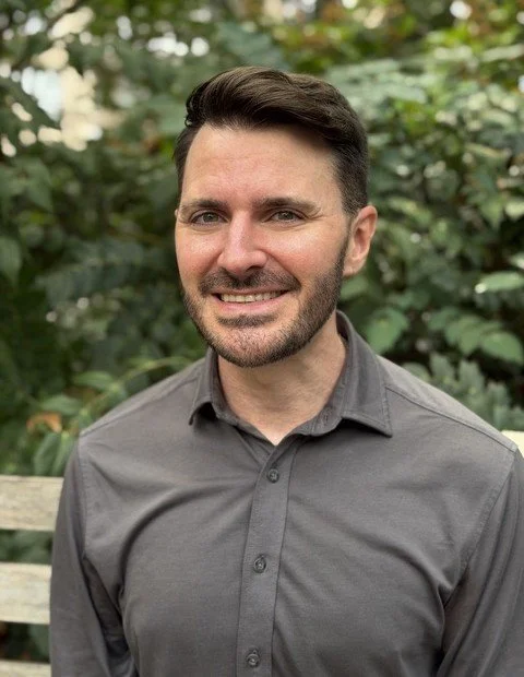 Headshot of Brian Higginbotham, LMSW, therapist at Upstate Counseling, smiling with short dark hair and a trimmed beard, wearing a gray button-down shirt, photographed outdoors with a softly blurred leafy green background.