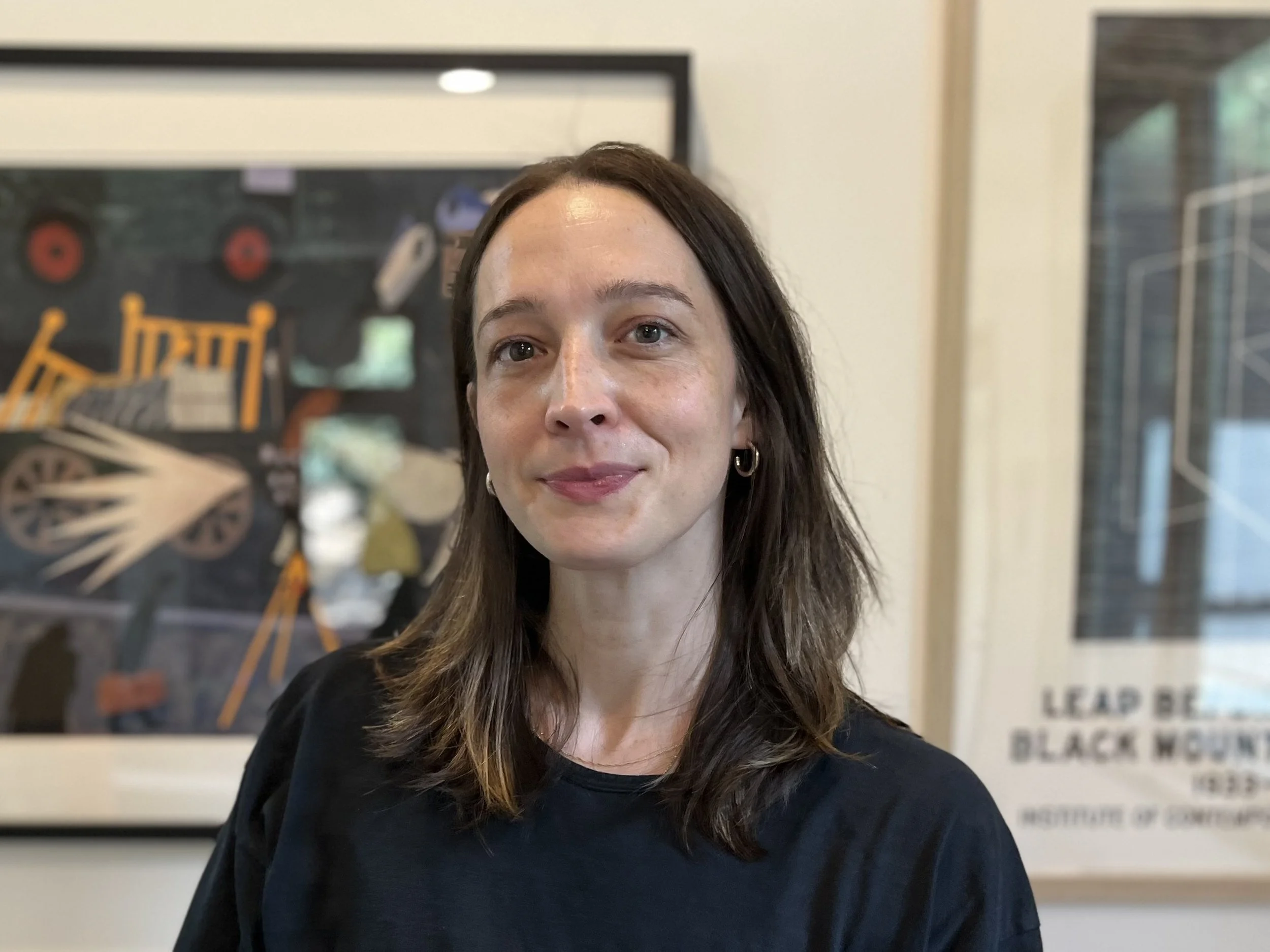 Headshot of Margaret Green, LMSW, therapist at Upstate Counseling, with shoulder-length brown hair, wearing small hoop earrings and a black top, standing indoors with framed artwork in the background.