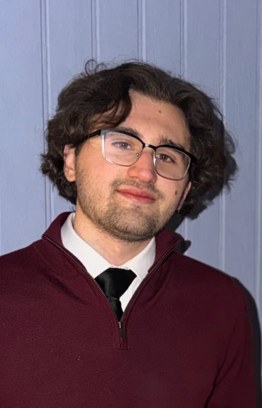 Headshot of Jack Bruce, Marketing & Design Coordinator at Upstate Counseling, with medium-length wavy dark hair and glasses, wearing a maroon quarter-zip sweater over a white shirt and black tie, standing against a light paneled background.