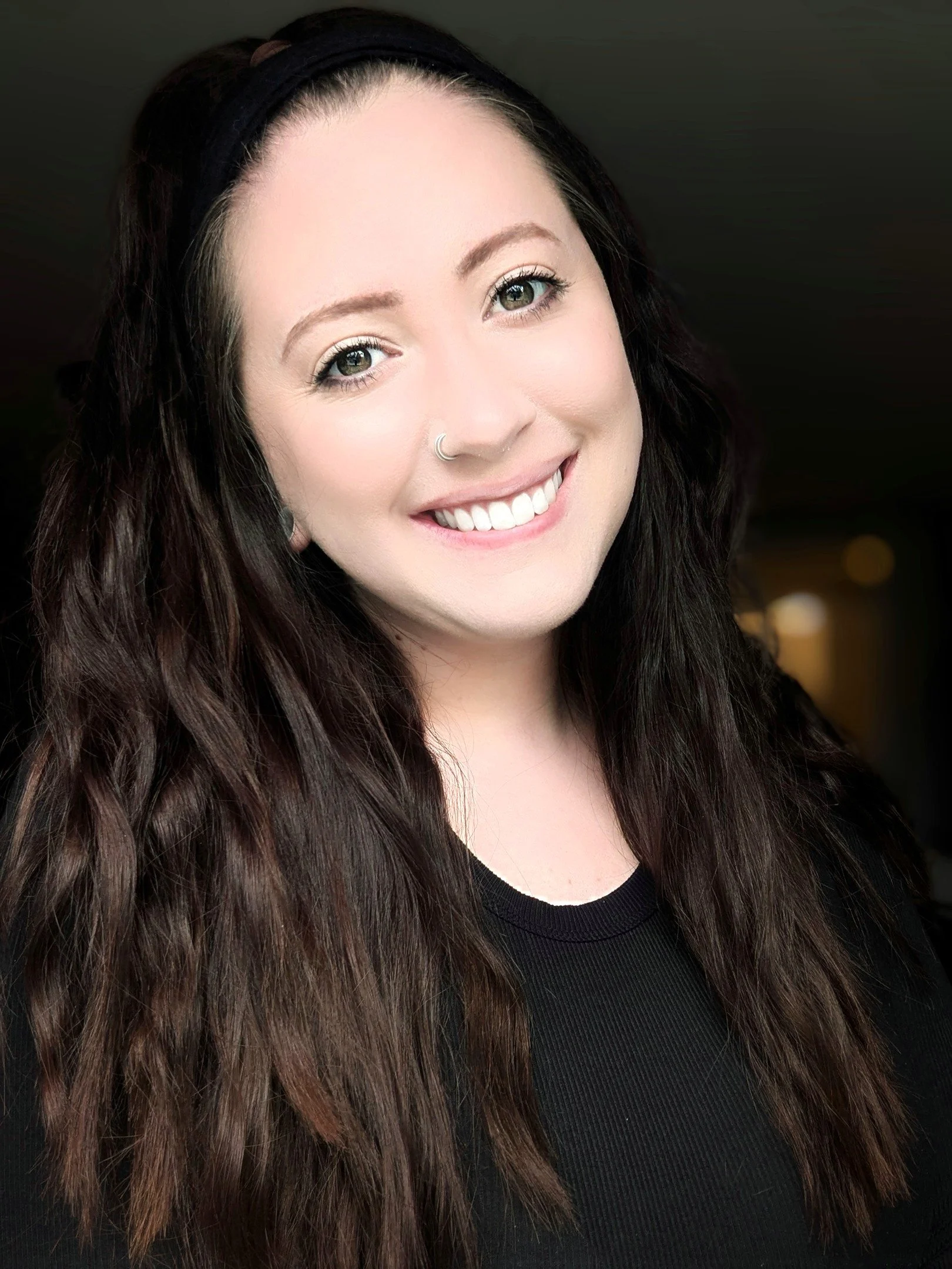 Headshot of Caitlyn Cramer, LMSW, therapist at Upstate Counseling, smiling with long dark wavy hair, wearing a black top and a nose ring, photographed indoors against a softly blurred dark background.
