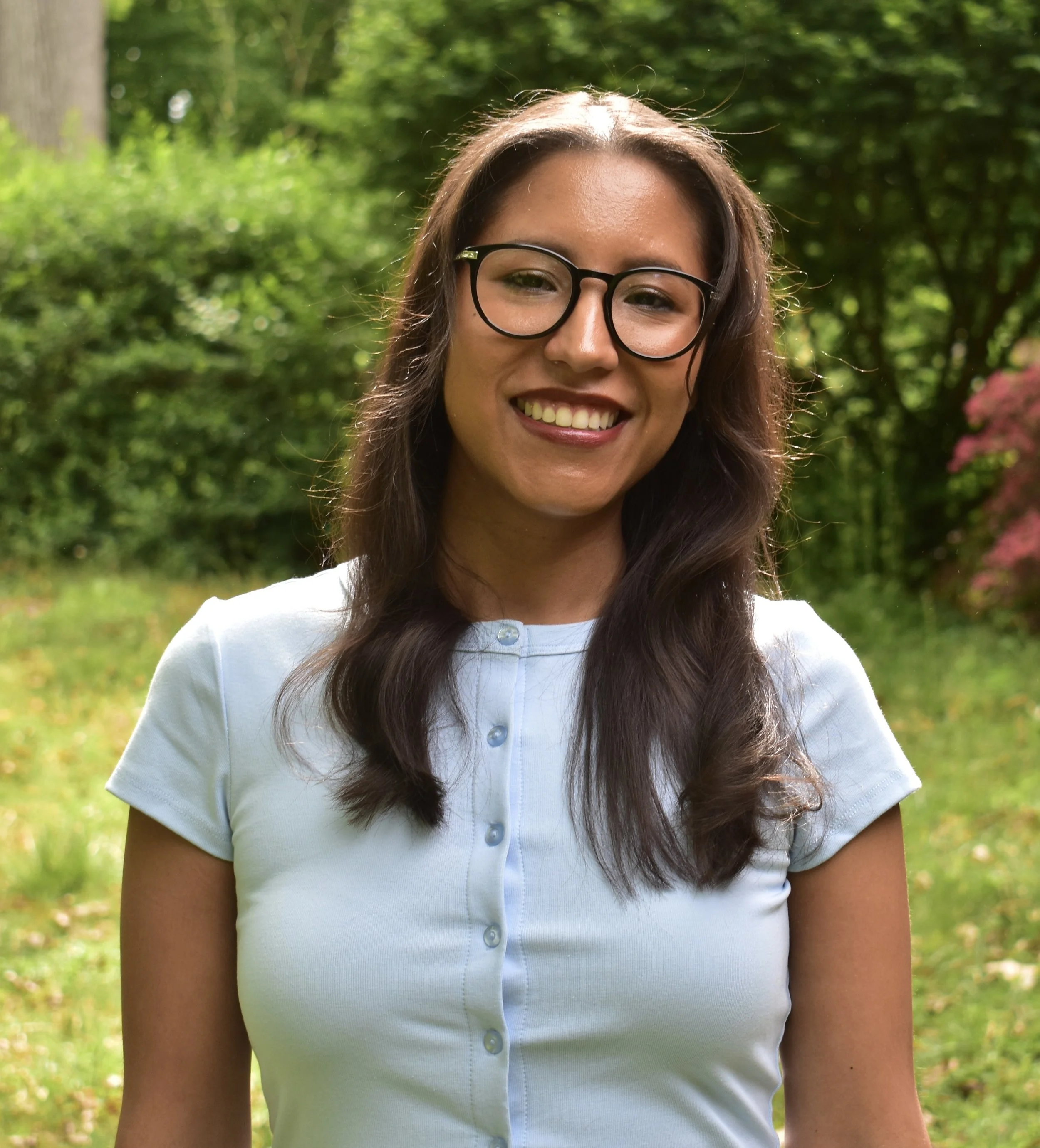 Headshot of Claire Burnham, LCSW, Clinical Supervisor at Upstate Counseling, smiling with long dark hair and glasses, wearing a light blue short-sleeve top, photographed outdoors with a softly blurred green natural background.