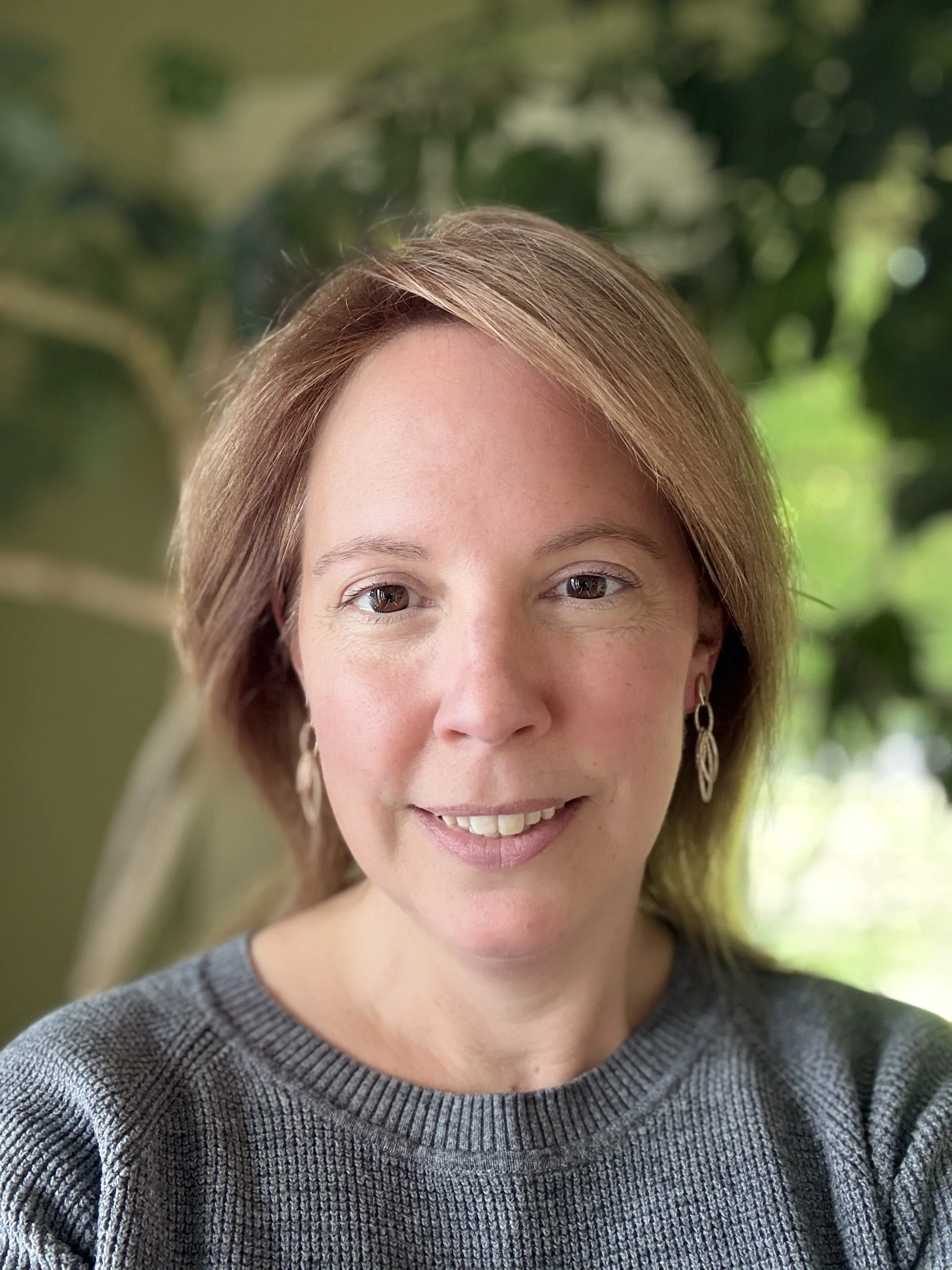 Headshot of Zoe Mangold, therapist at Upstate Counseling, with light brown hair worn down, wearing drop earrings and a gray knit sweater, photographed indoors with a softly blurred leafy background.