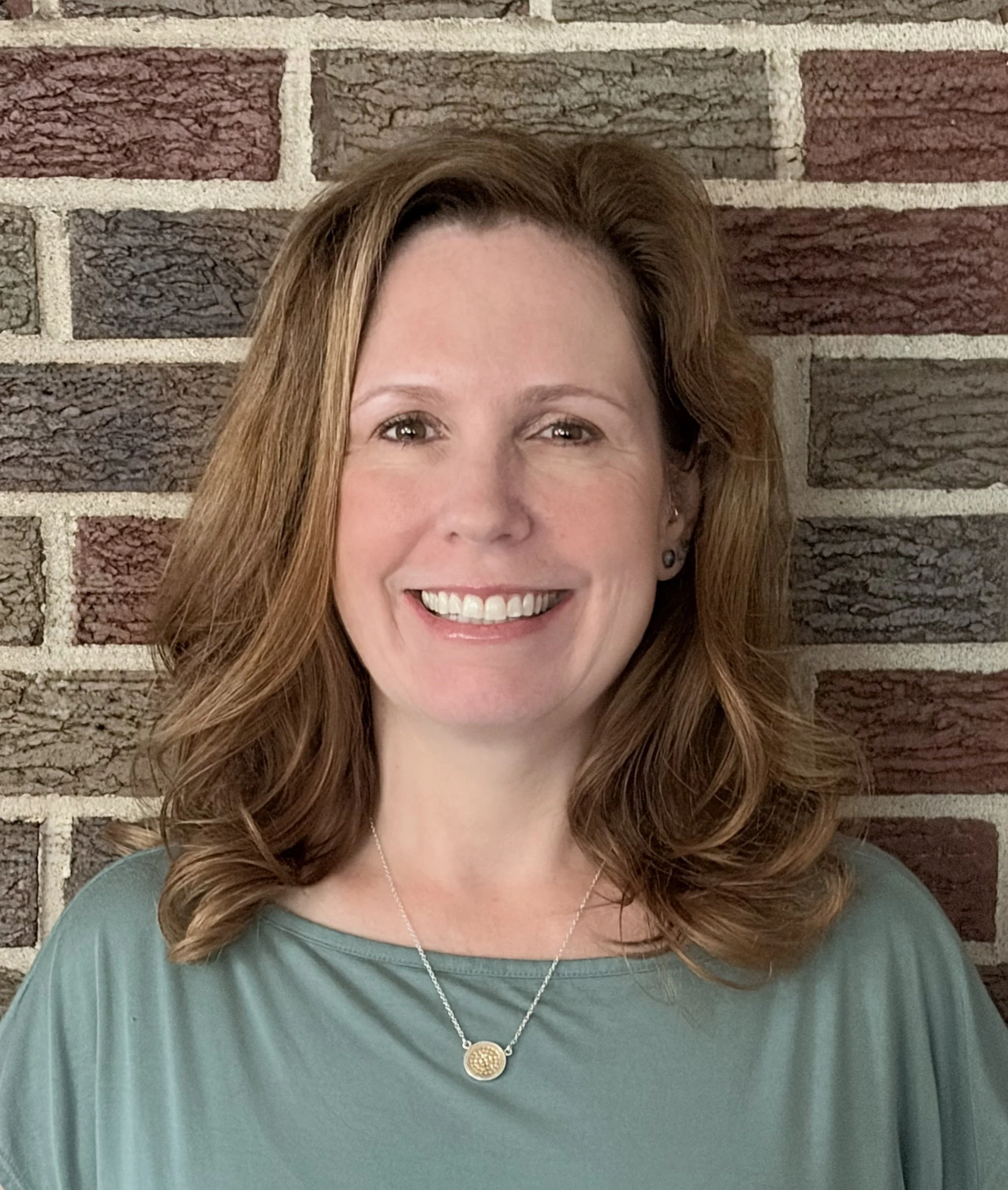 Headshot of Jennifer Barry, LMSW, therapist at Upstate Counseling, smiling with shoulder-length wavy light brown hair, wearing a sage green top and pendant necklace, photographed against a textured brick background.
