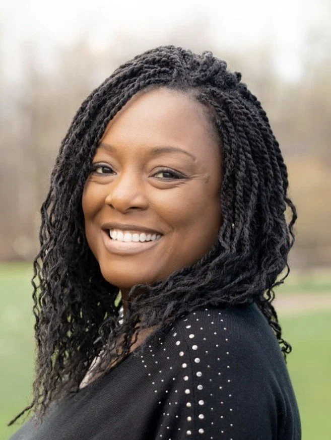 Headshot of Bree Taylor-Farnsworth, LMSW, therapist at Upstate Counseling, smiling with shoulder-length twisted hair, wearing a black top with subtle stud detailing, photographed outdoors with a softly blurred natural background.
